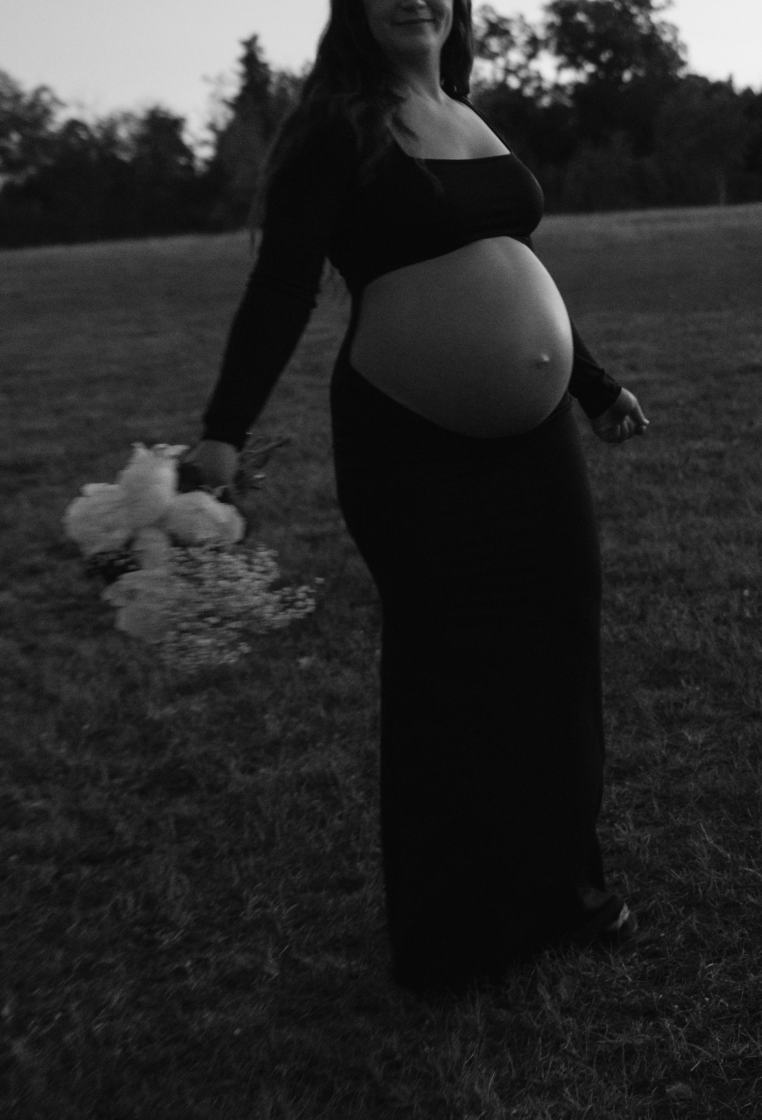 Black and white maternity photo of pregnant woman in black dress holding white flowers in a field.
