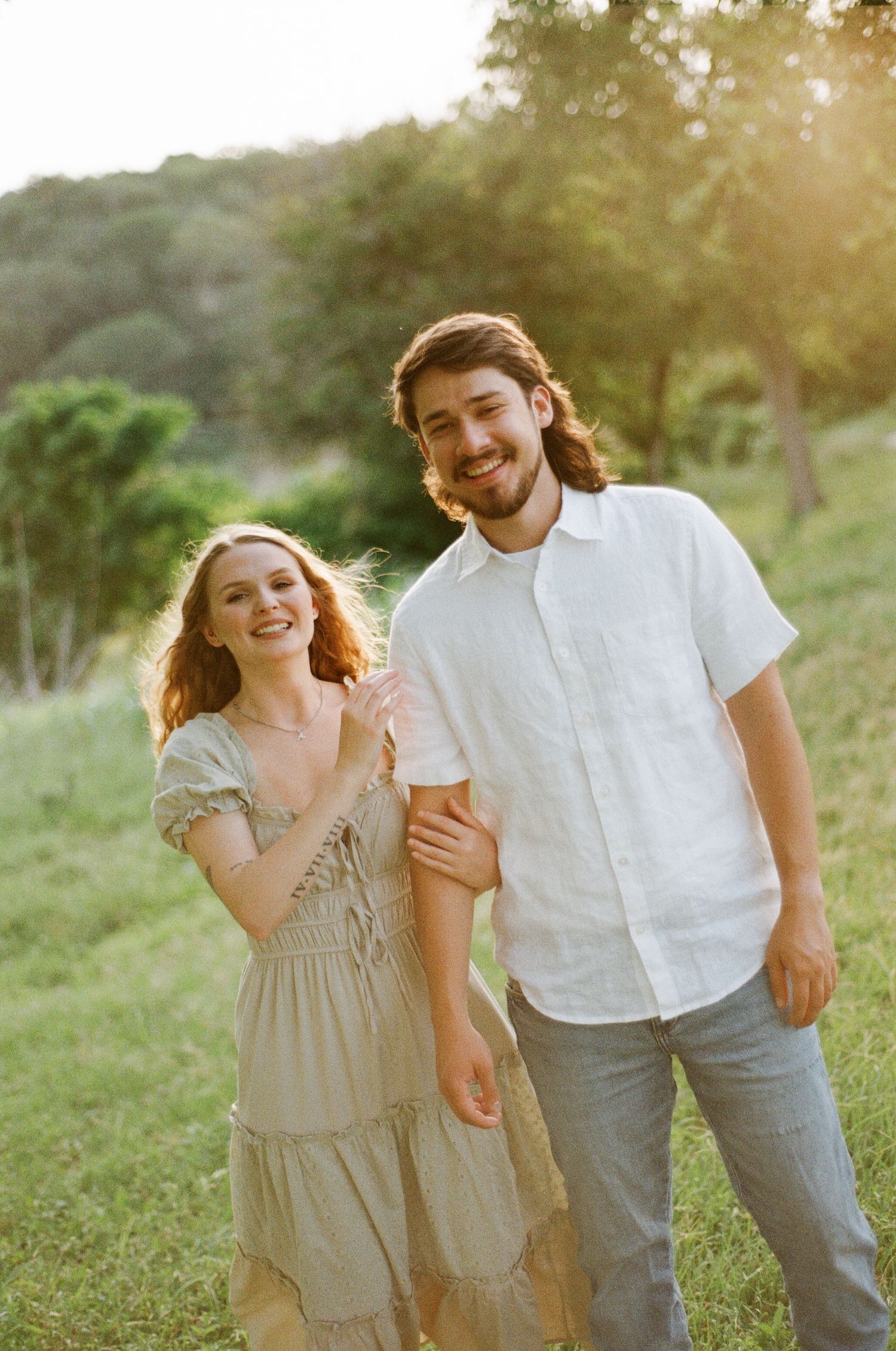 A smiling couple poses outdoors in golden hour light, woman in flowy dress, man in white linen shirt on green meadow.