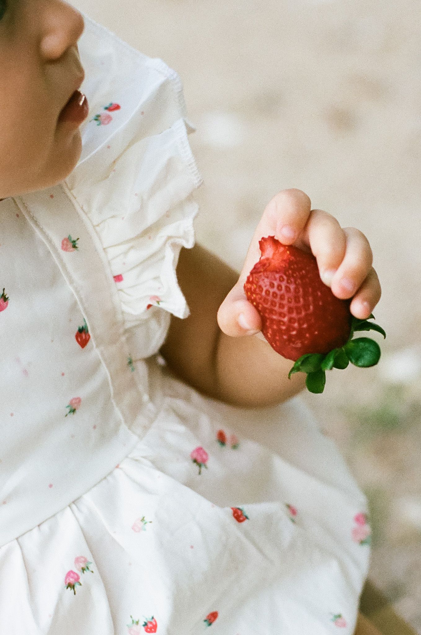 Young child in white strawberry-print dress holding a large fresh red strawberry with green leaves.