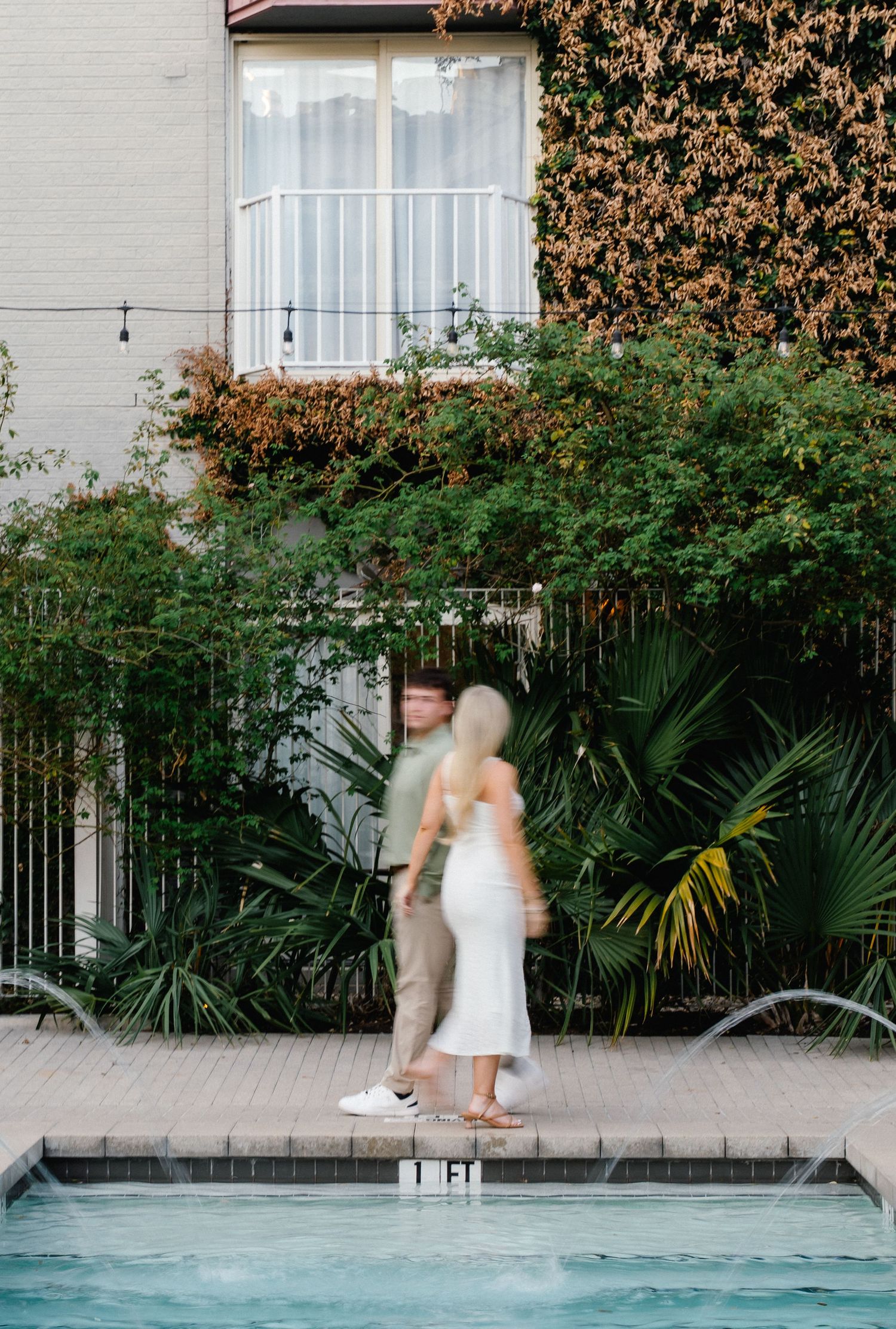 Couple embracing near a pool with lush tropical greenery and ivy-covered building backdrop.