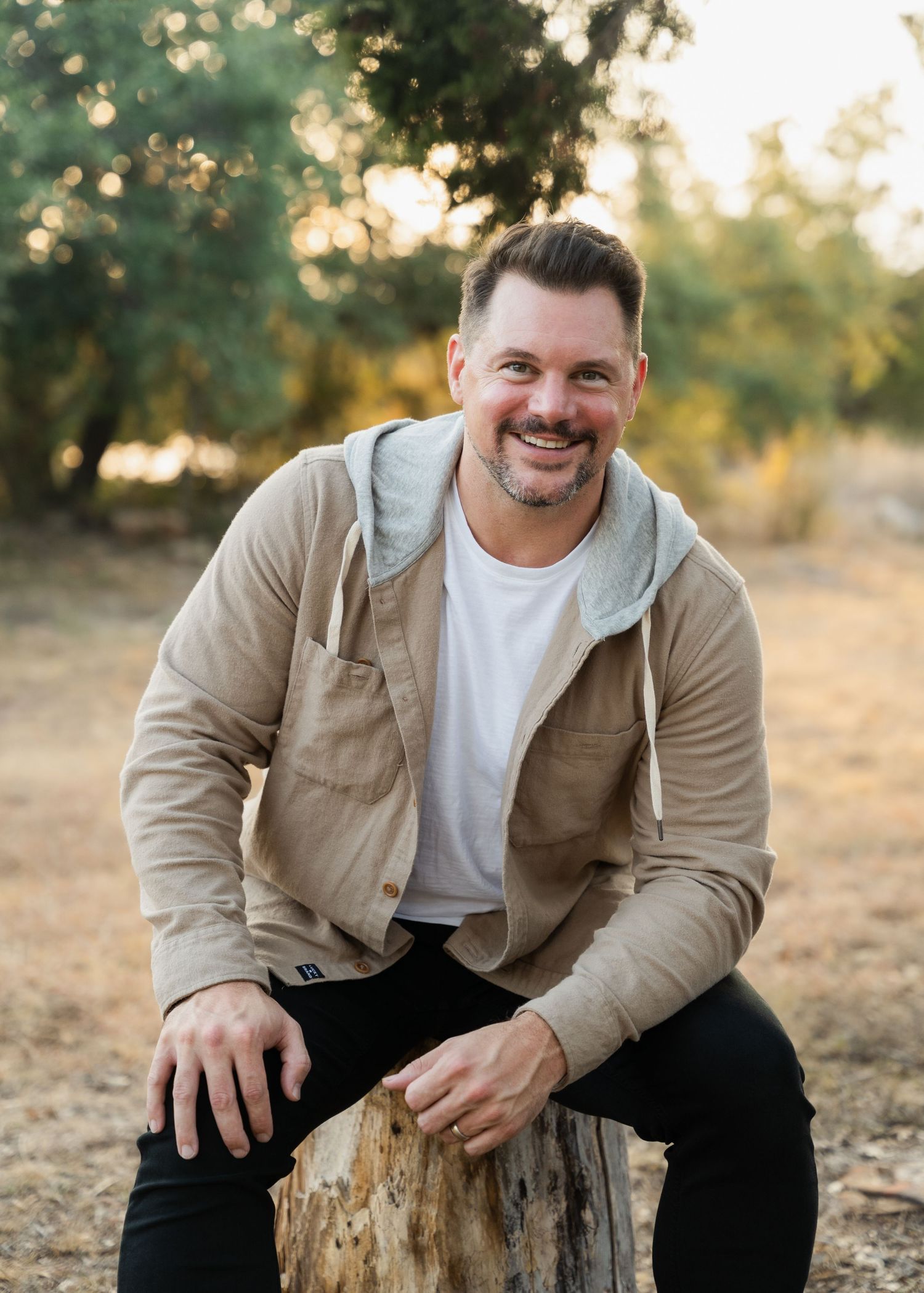 Smiling man in beige hoodie jacket sitting on tree stump in sunny outdoor setting with trees in background.