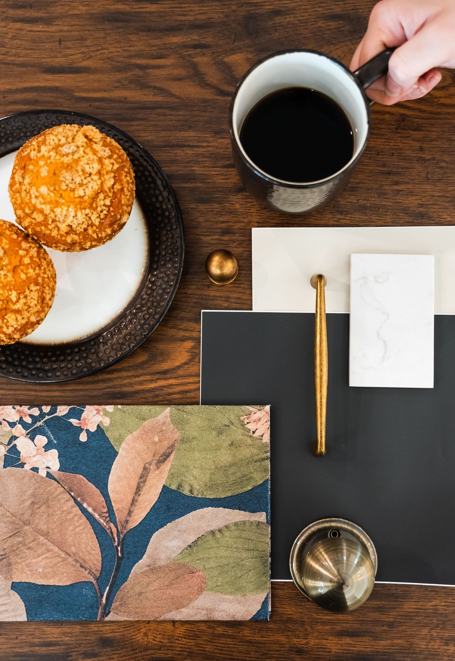 Overhead view of coffee, muffins, design samples, and gold stationery on a dark wood table.
