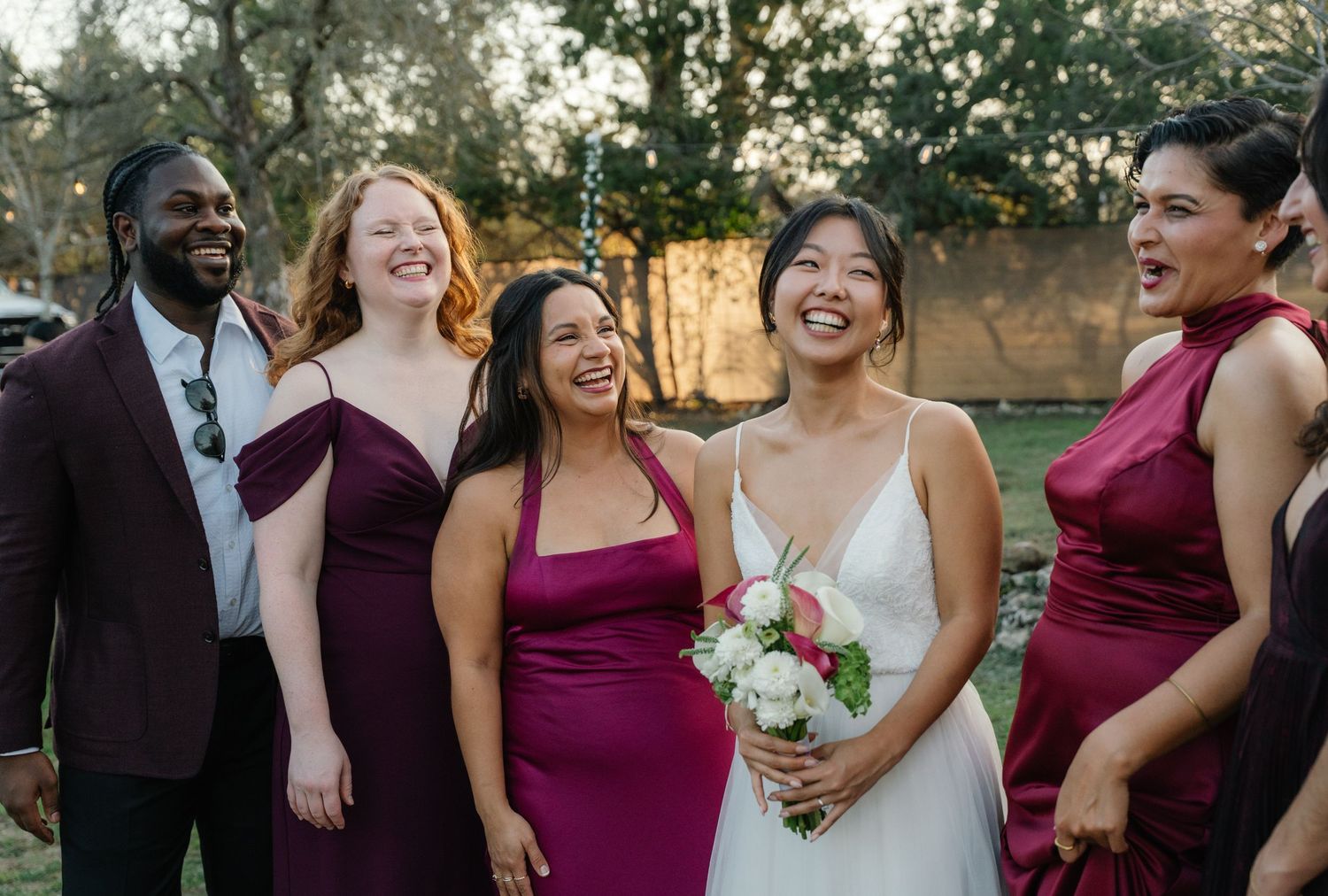 Bride laughing with diverse bridesmaids in burgundy dresses at outdoor wedding.
