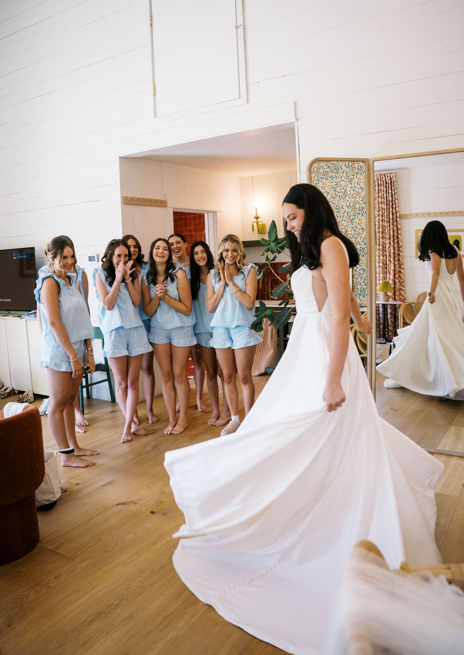 Bridesmaids in light blue matching sets react with joy as bride twirls white wedding gown in bright bridal suite.