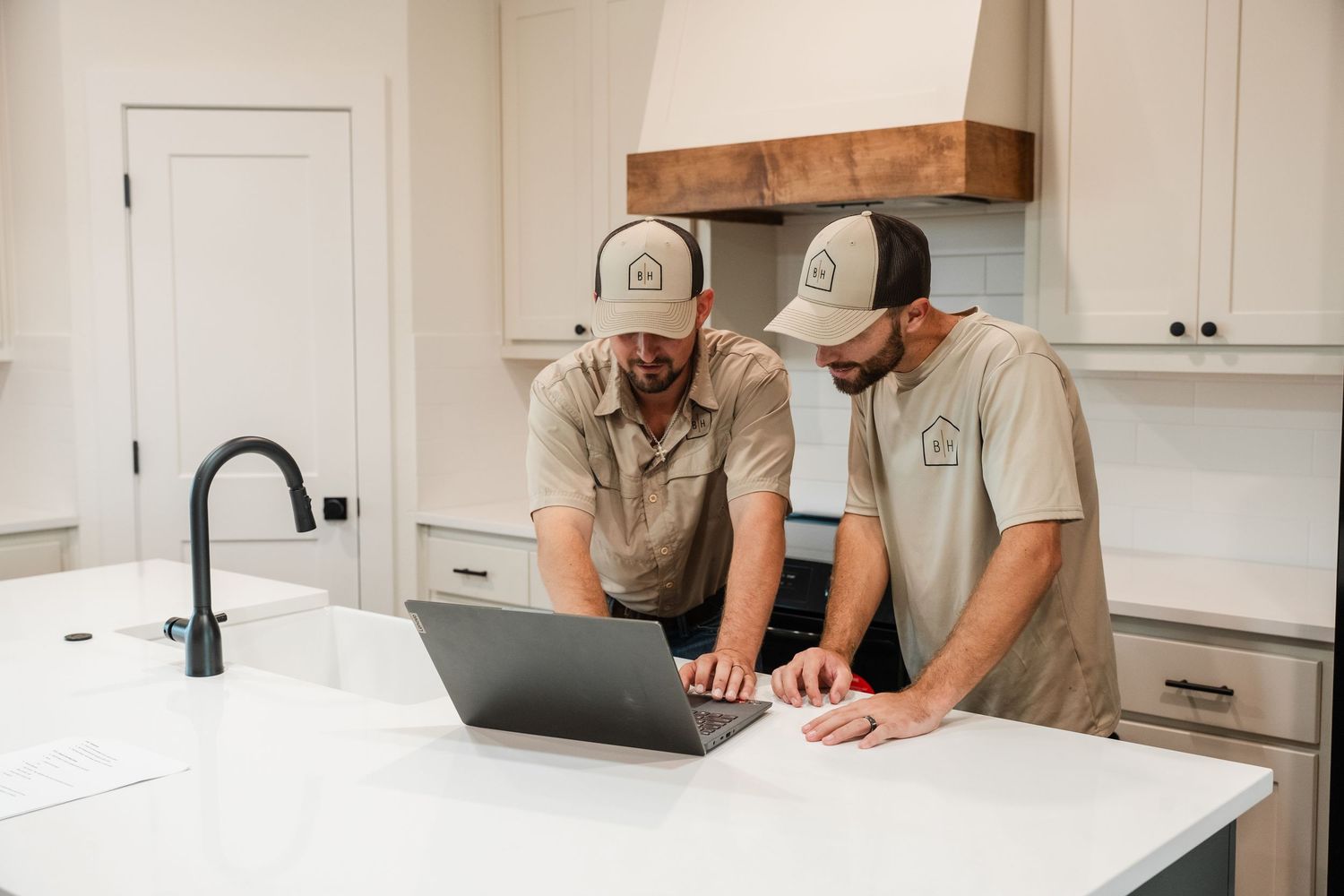 Two builders in matching caps reviewing plans on a laptop at a white kitchen island.
