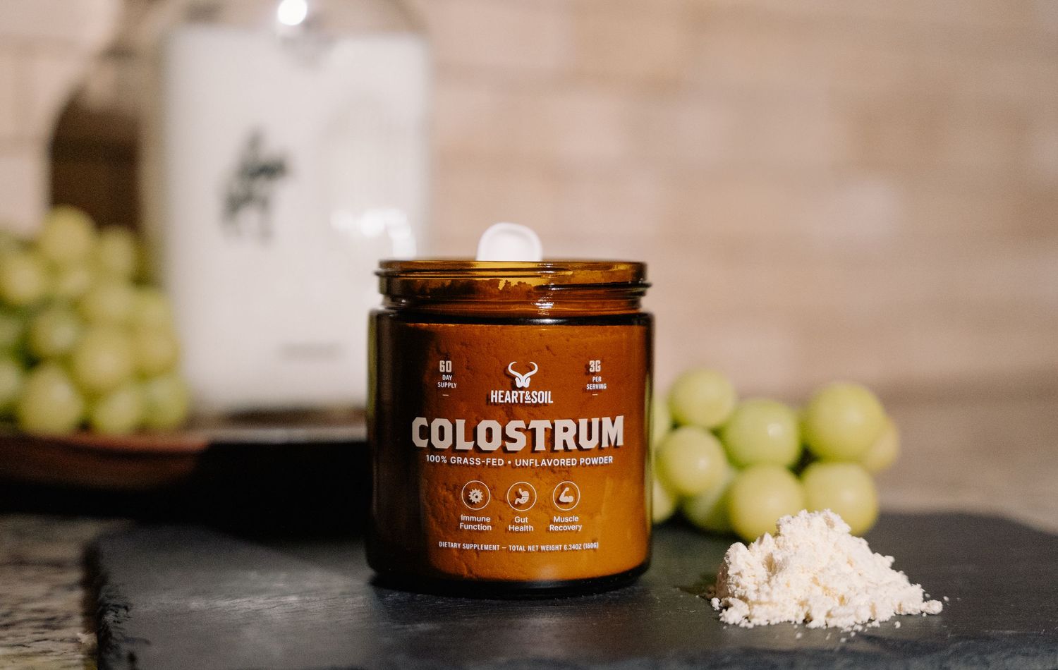 Amber jar of colostrum powder supplement on slate board with green grapes and white powder sample nearby.