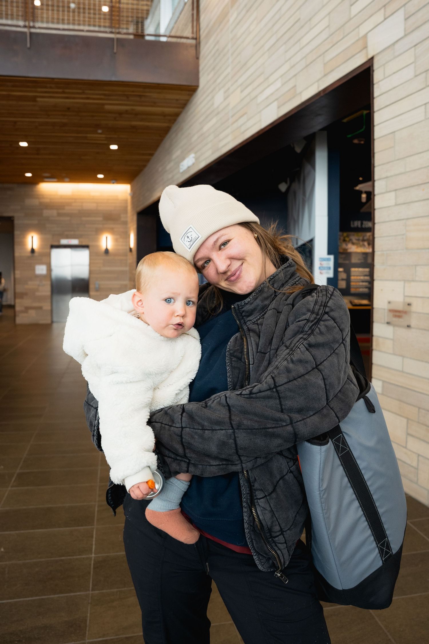 Smiling woman in winter hat holding a baby in white jacket inside modern building.