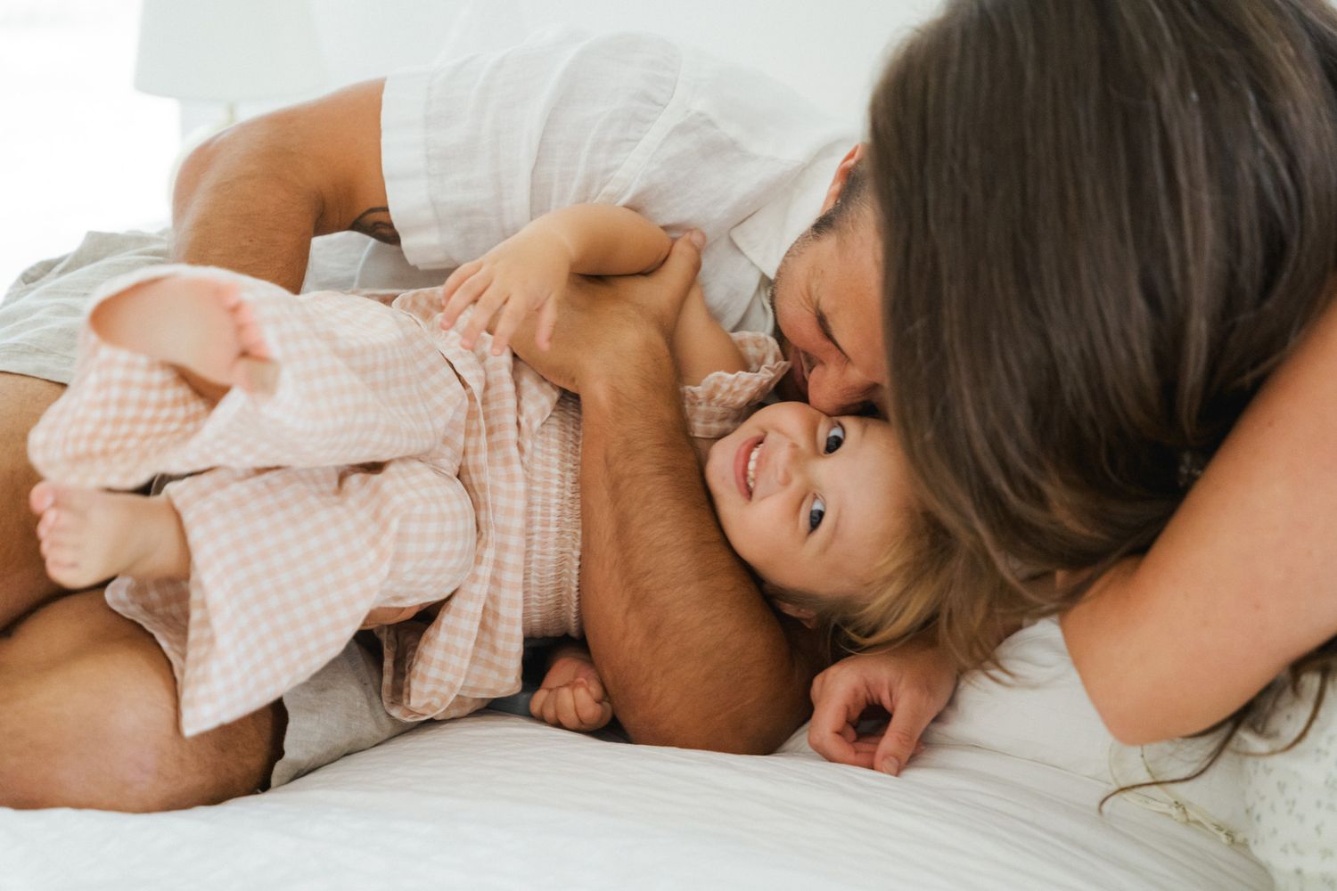A mother tenderly kisses her laughing baby while lying together on a white bed.