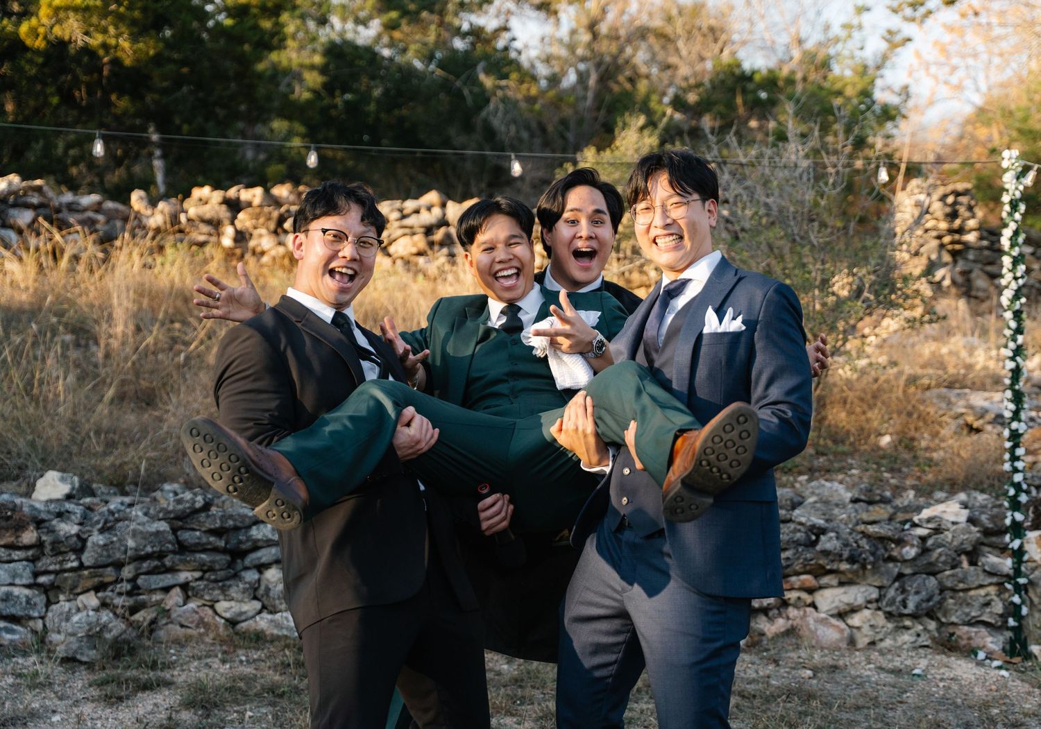 Groom and groomsmen laughing and lifting each other at outdoor wedding celebration.