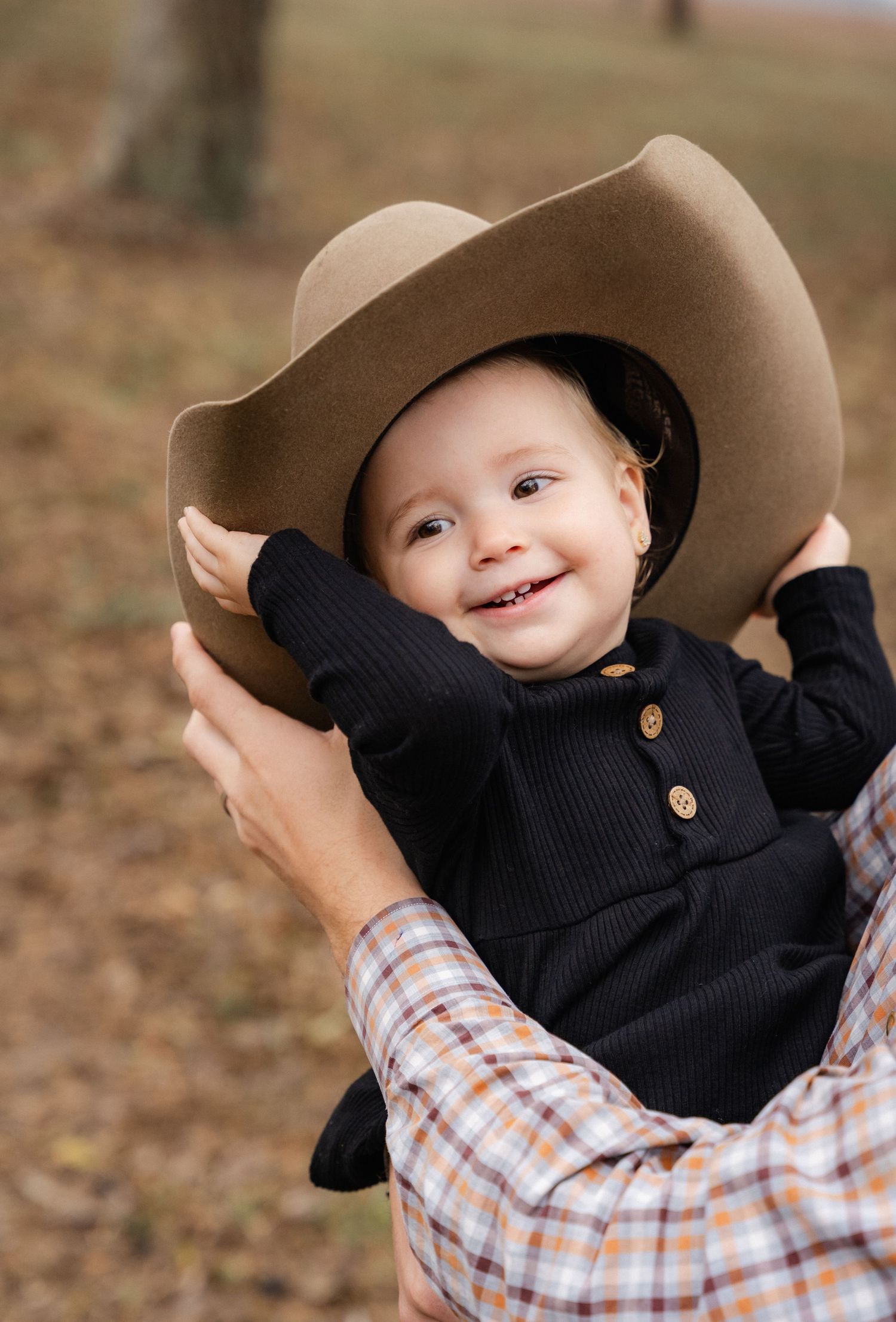 Adorable smiling baby wearing an oversized tan cowboy hat being held outdoors in autumn.