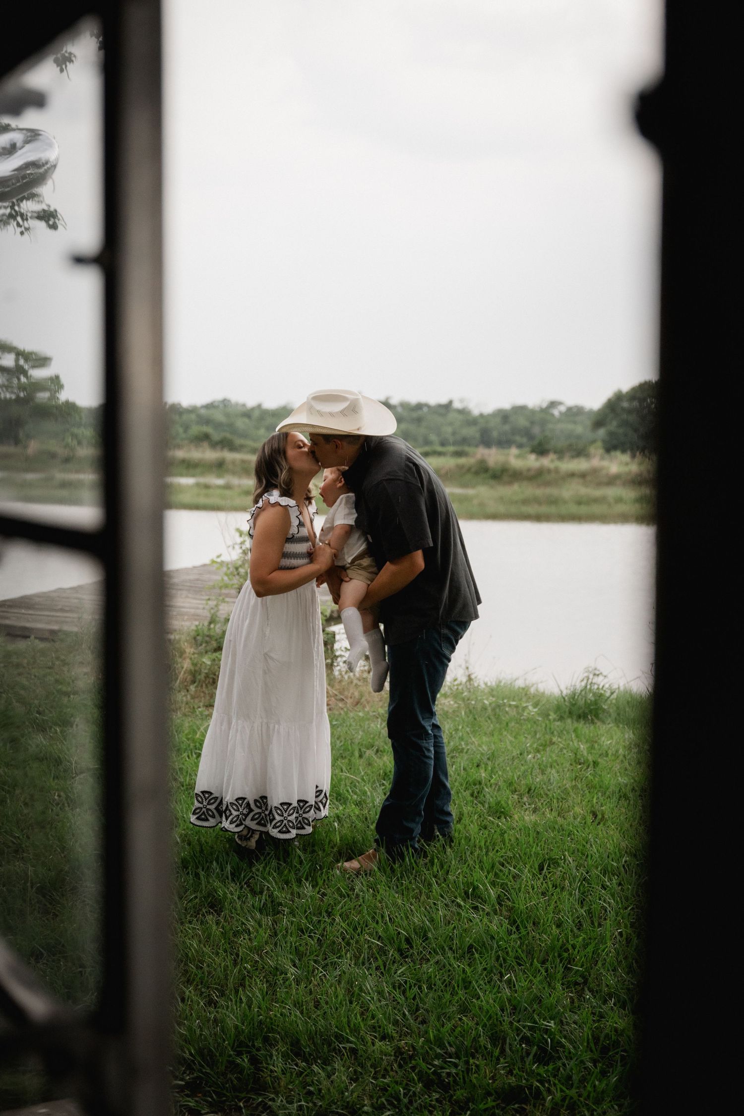 Couple sharing a romantic kiss while holding their baby near a serene lake on a farm.