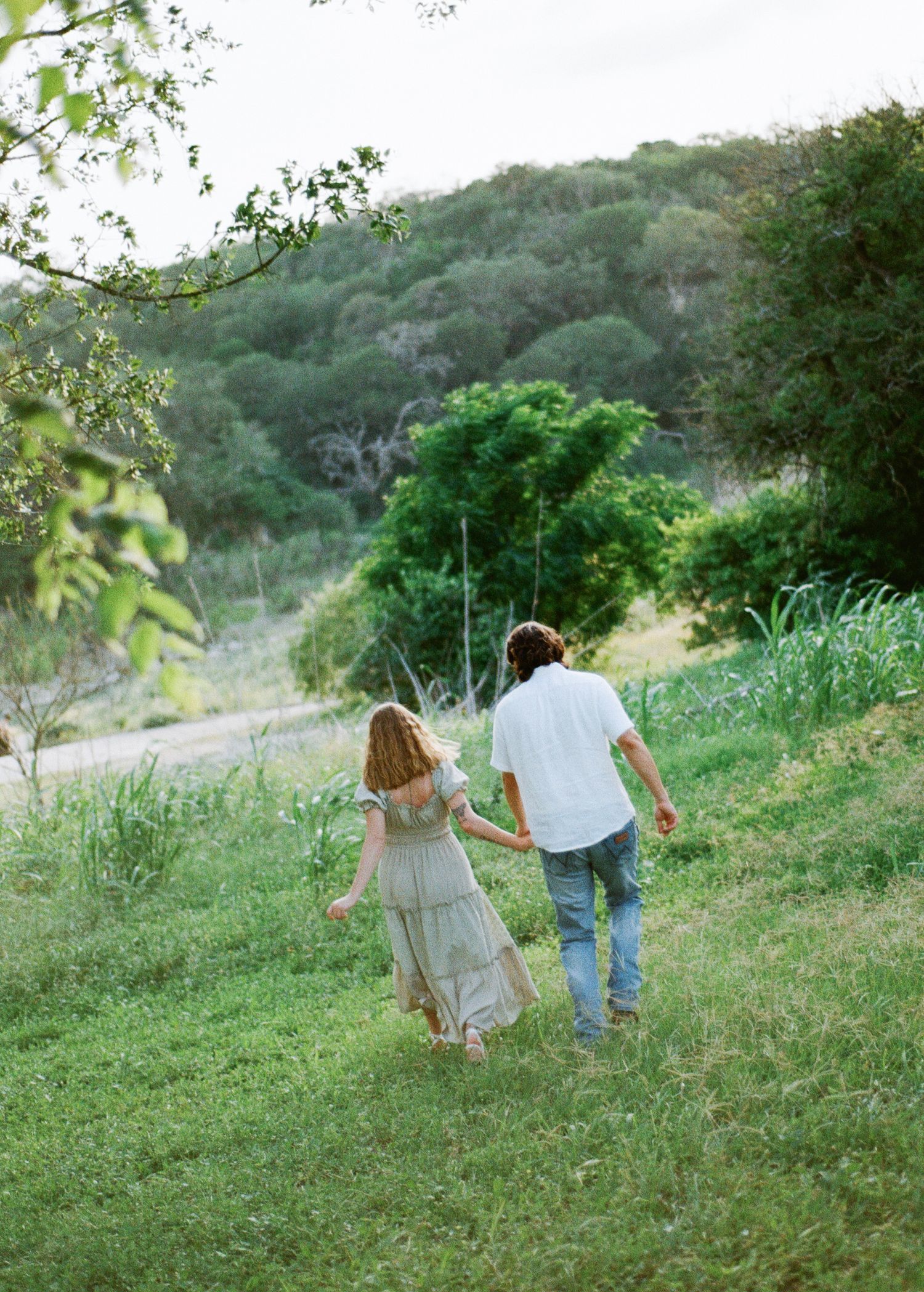 Couple holding hands walking through lush green meadow with rolling hills and trees in background.