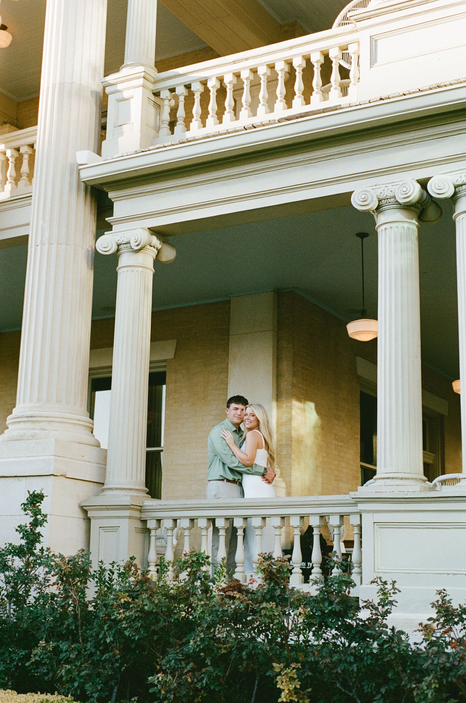 Happy couple embracing on the porch of a grand white columned Southern mansion at golden hour.