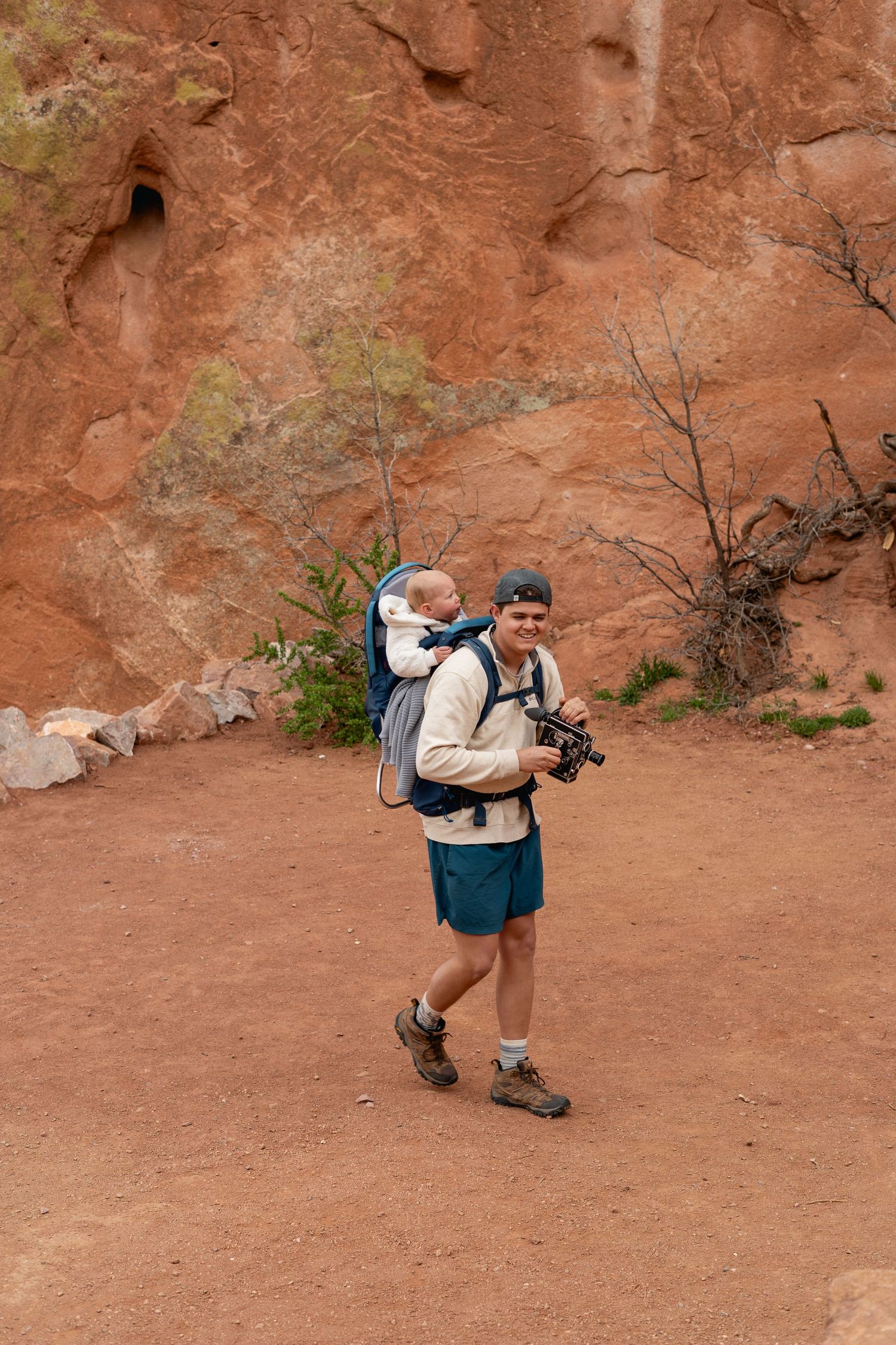 Smiling hiker carries baby in backpack carrier while holding camera near red rock formations outdoors.