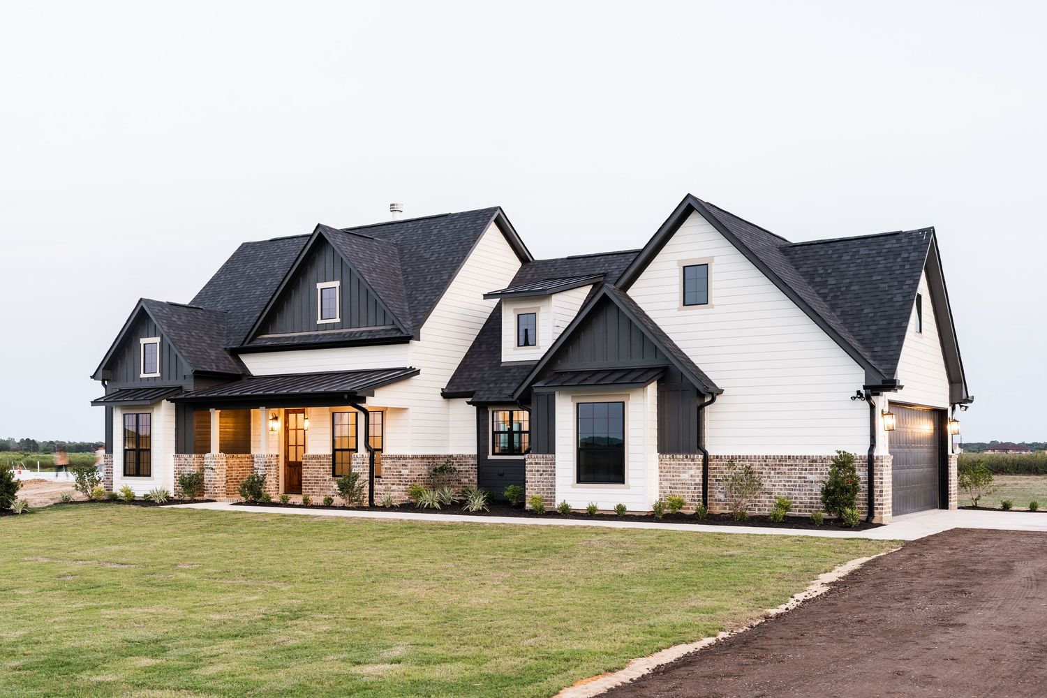 Elegant white farmhouse with black roof, stone facade, board and batten siding at dusk.