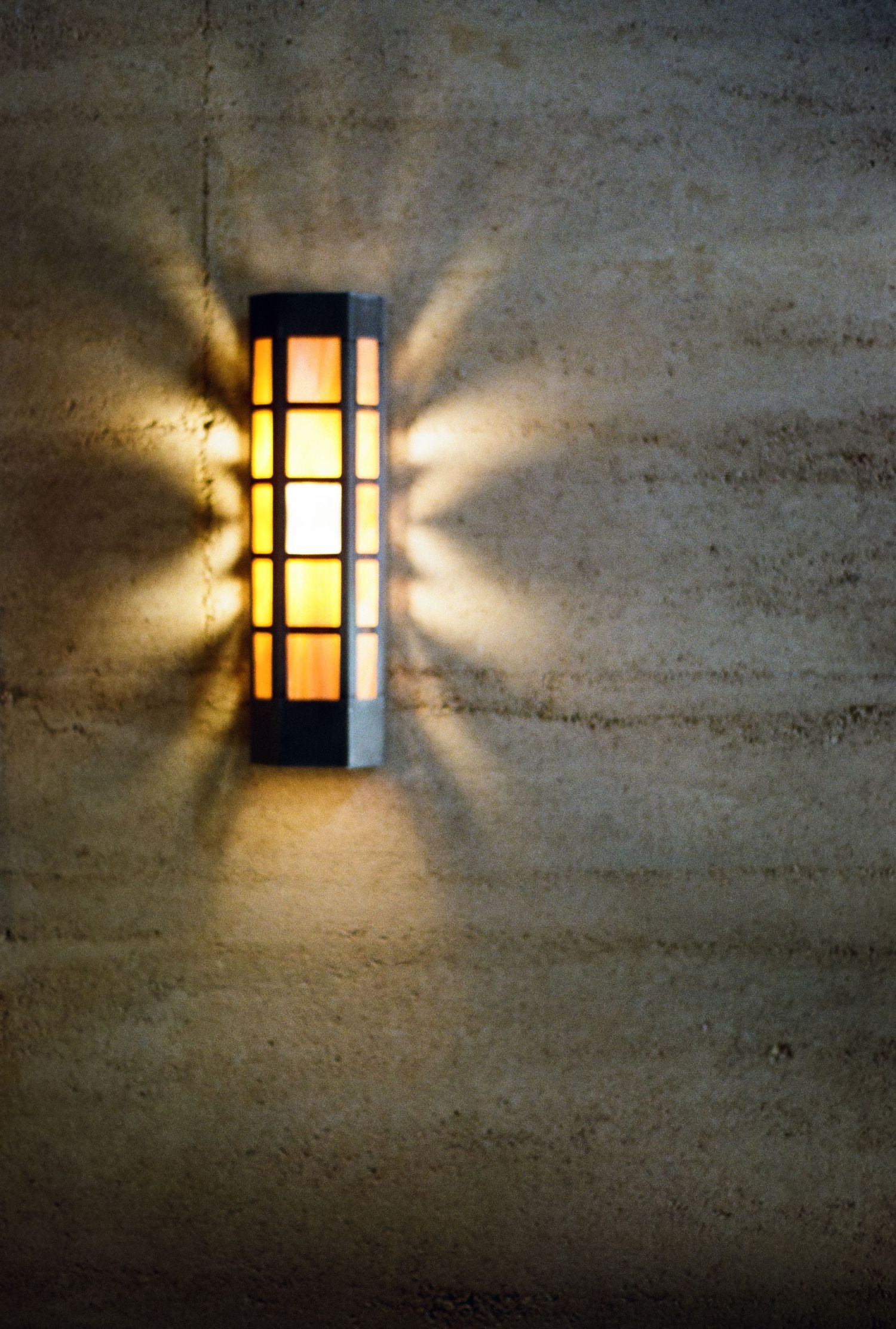 Glowing rectangular wall sconce casting dramatic symmetrical light rays on a textured limestone exterior wall at night.