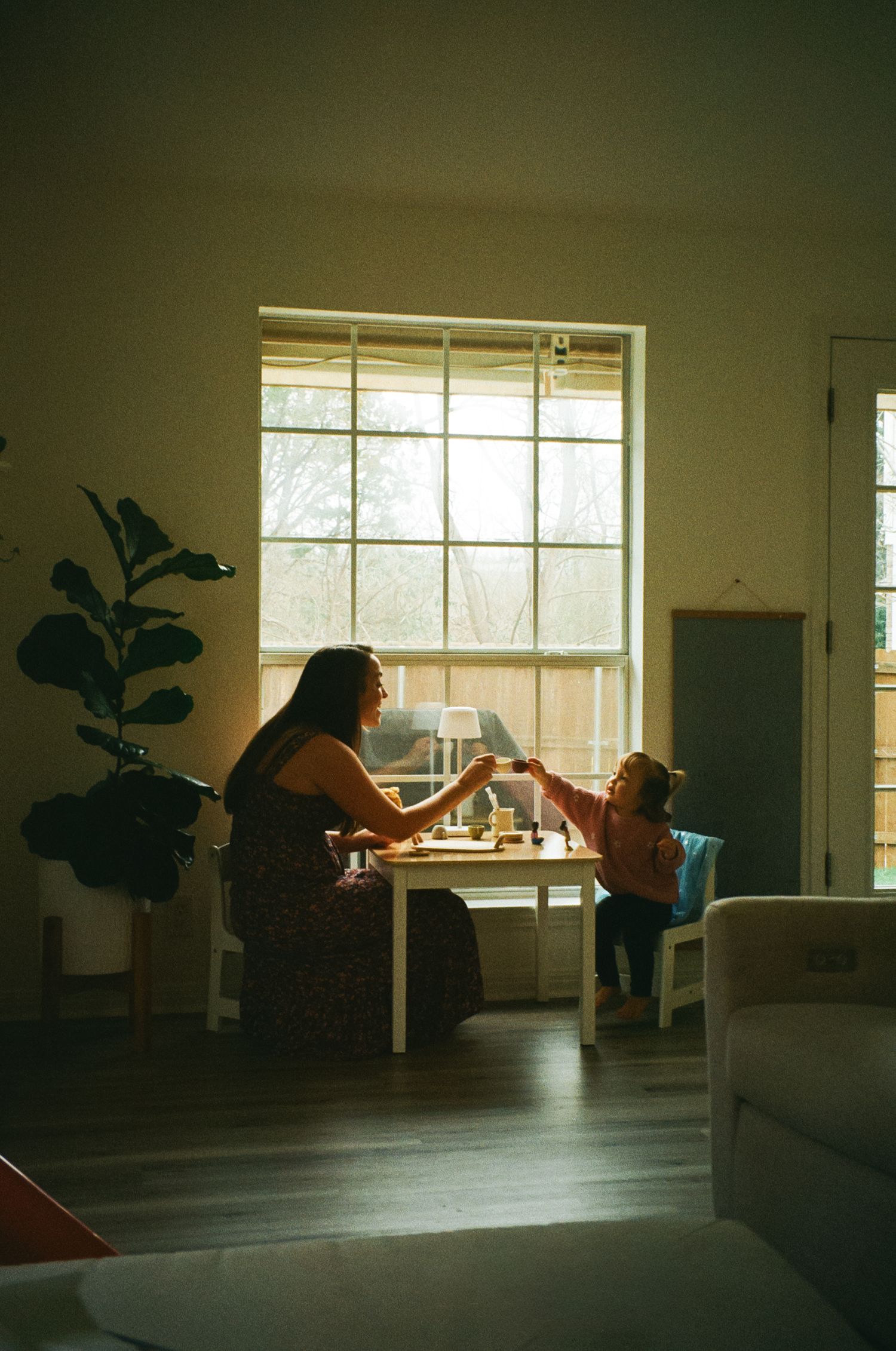 A mother and toddler share a warm moment at a small table by a sunlit window indoors.