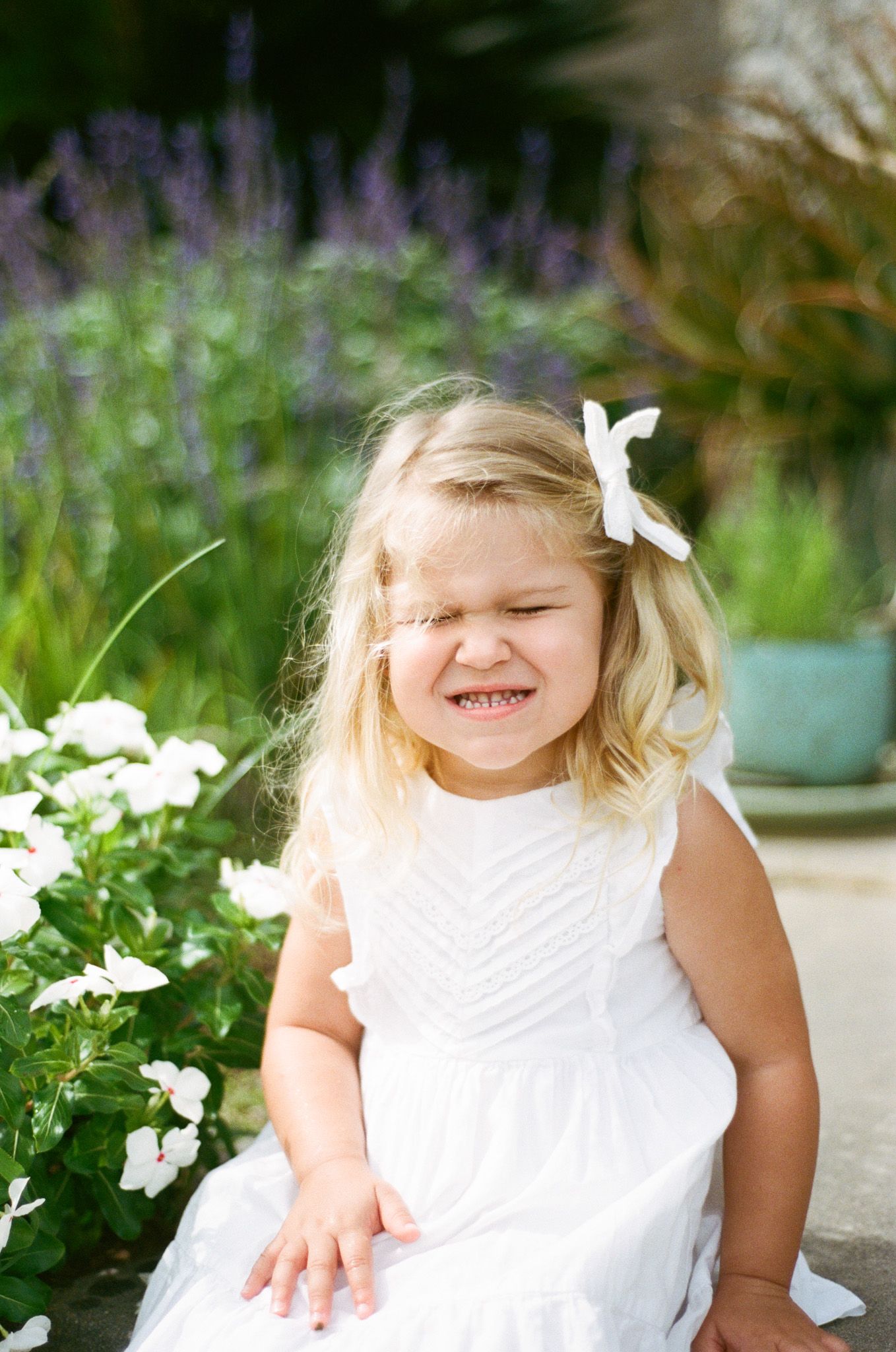An adorable blonde toddler girl in a white dress squints and grins beside white flowers in a garden.