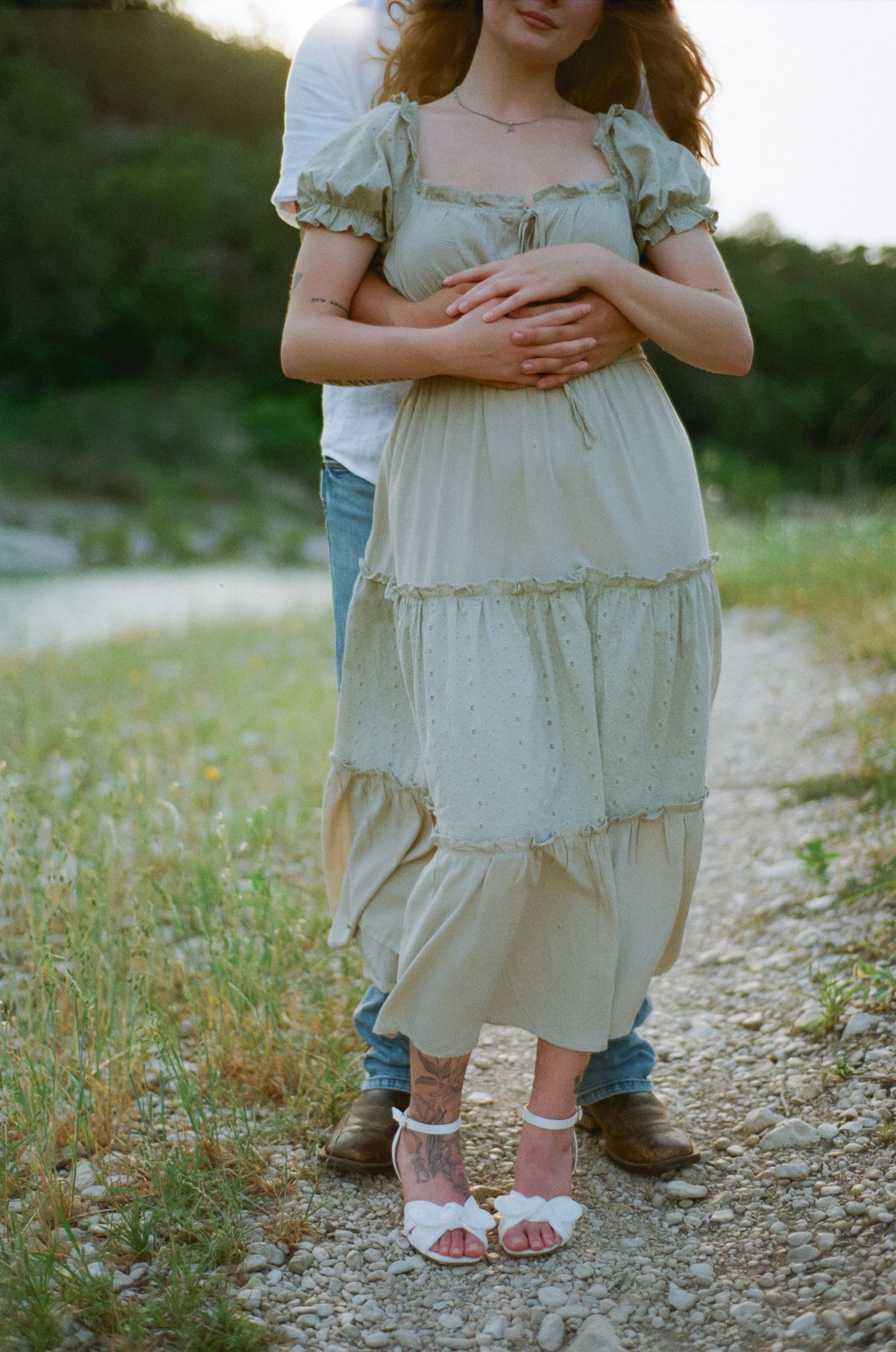 Couple embracing outdoors, woman in tiered sage green dress with white heels on a gravel path.