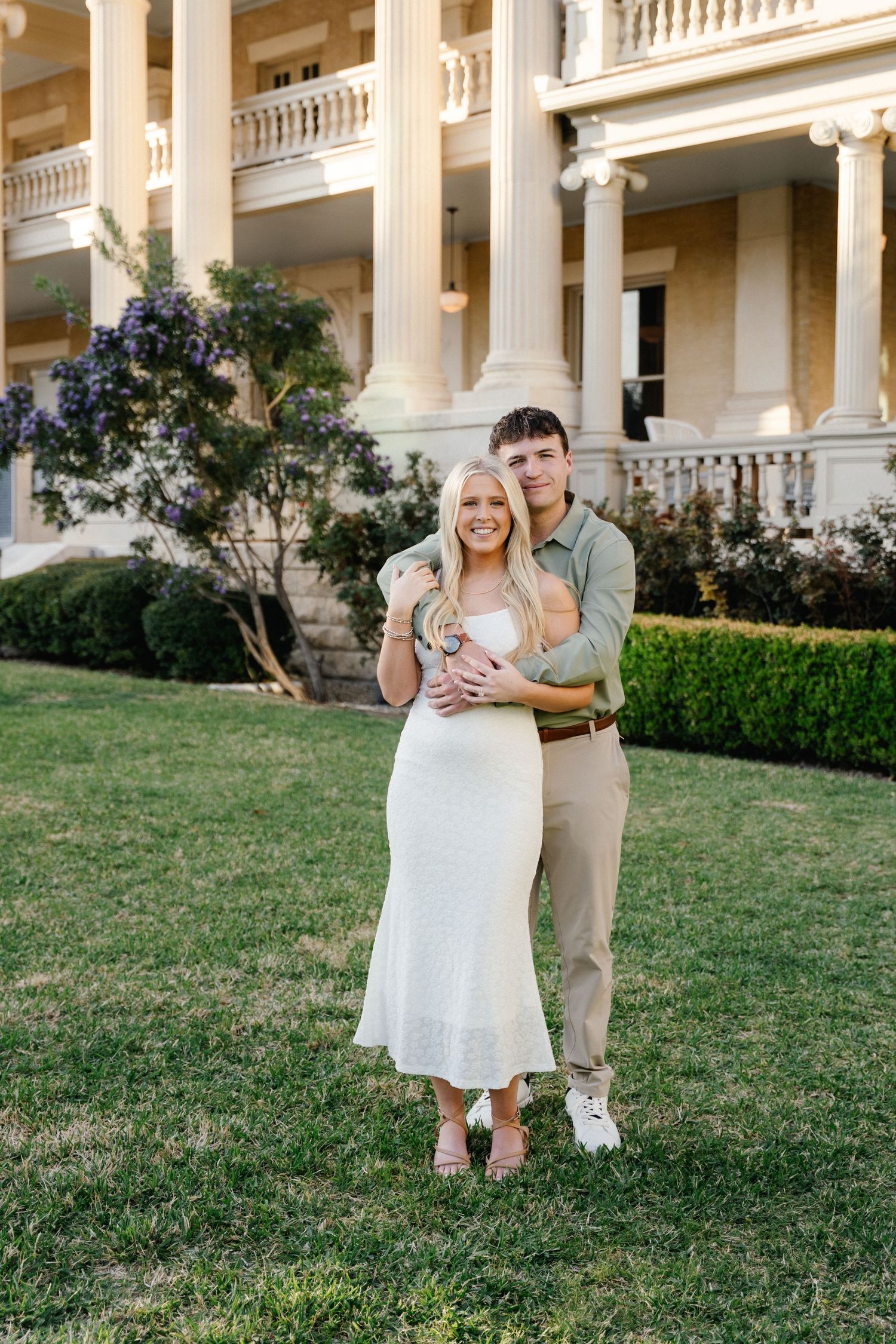 Couple posing in front of a grand white mansion with columns, woman in white dress, man in green shirt on green lawn.