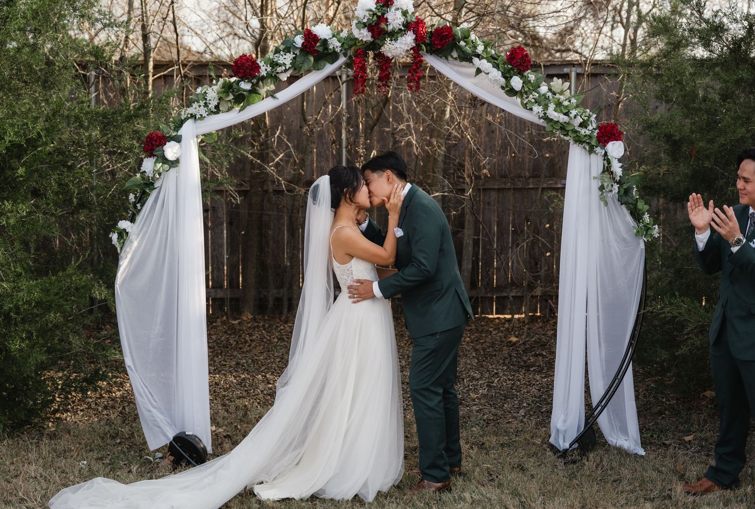 Bride and groom share first kiss under elegant white arch decorated with red and white roses.
