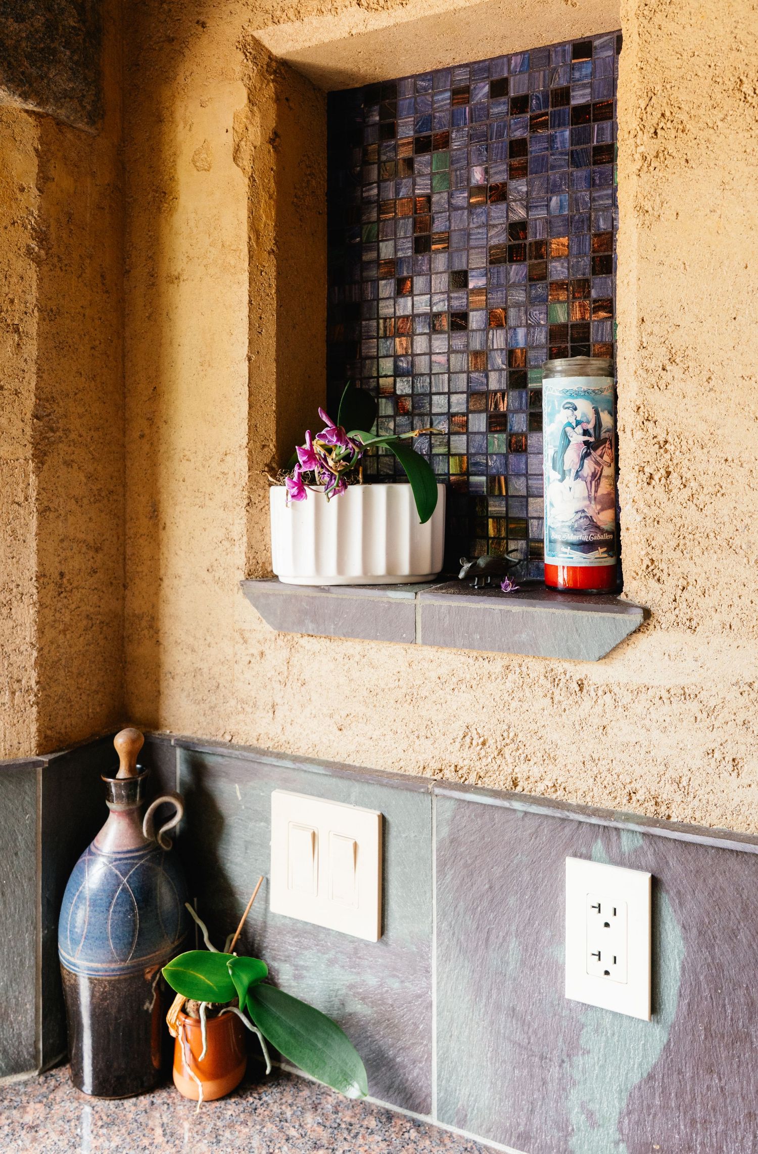 Recessed wall niche with iridescent mosaic tile backsplash displaying orchid plant and decorative candle on stone ledge.