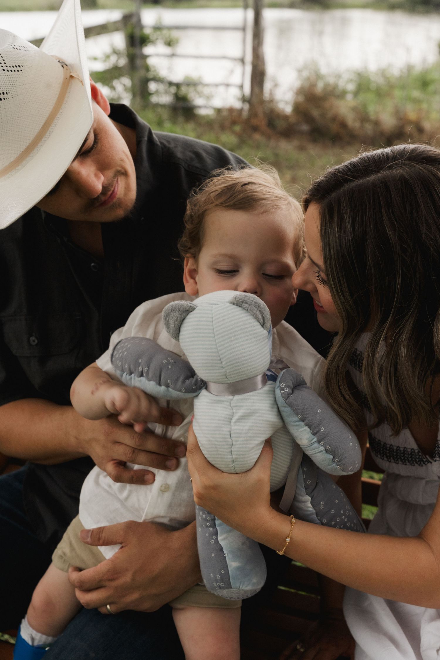 Parents lovingly lean in toward their toddler holding a gray elephant stuffed animal outdoors.