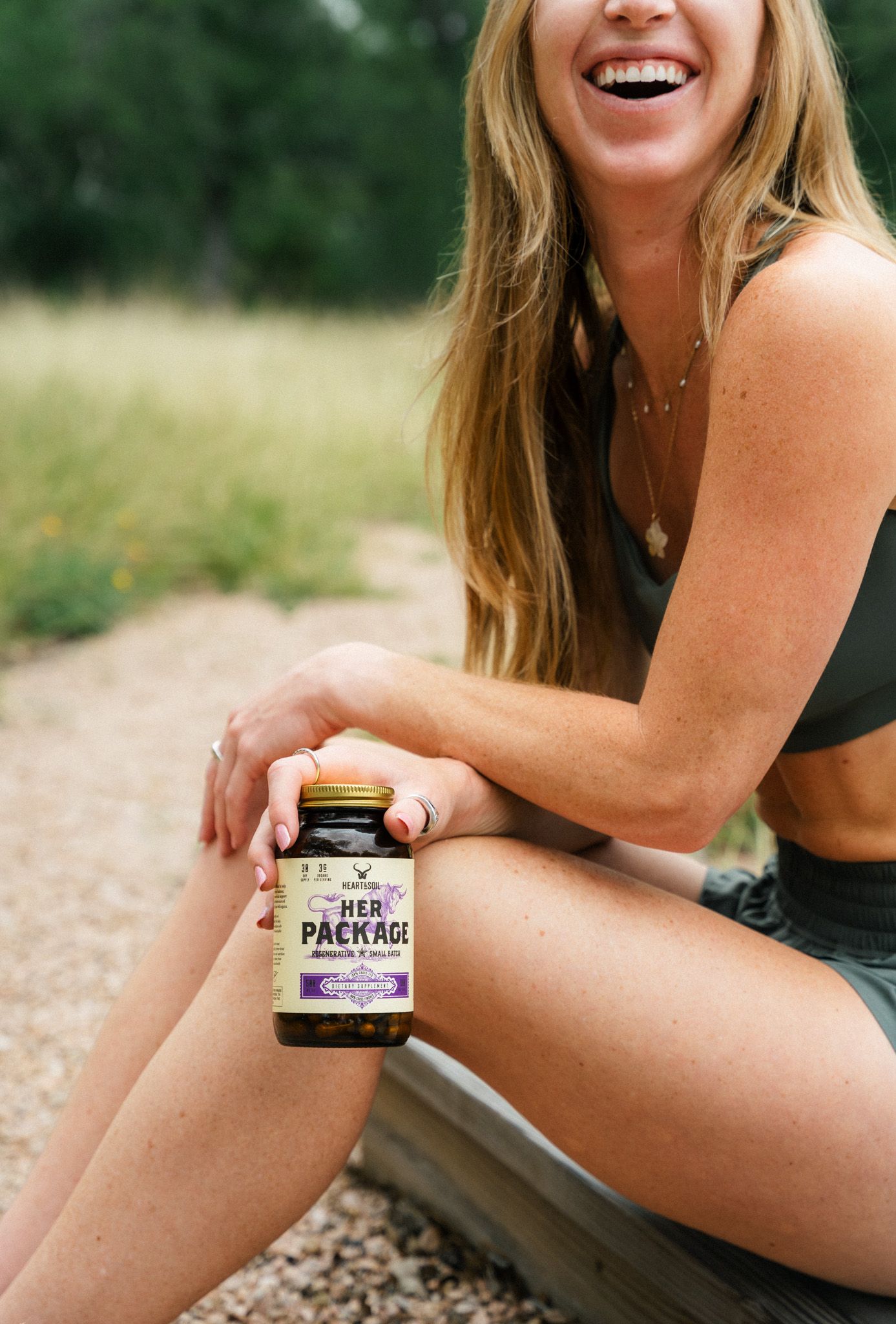 Smiling woman sitting outdoors holding a jar of Her Package supplements on a sunny day.