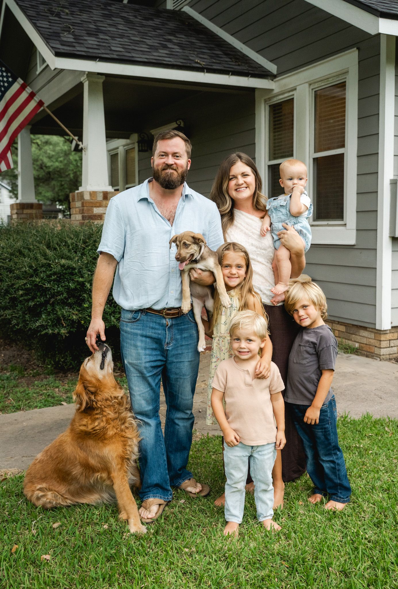 Happy family with two dogs poses in front of their home, including parents, three young children, and an American flag.