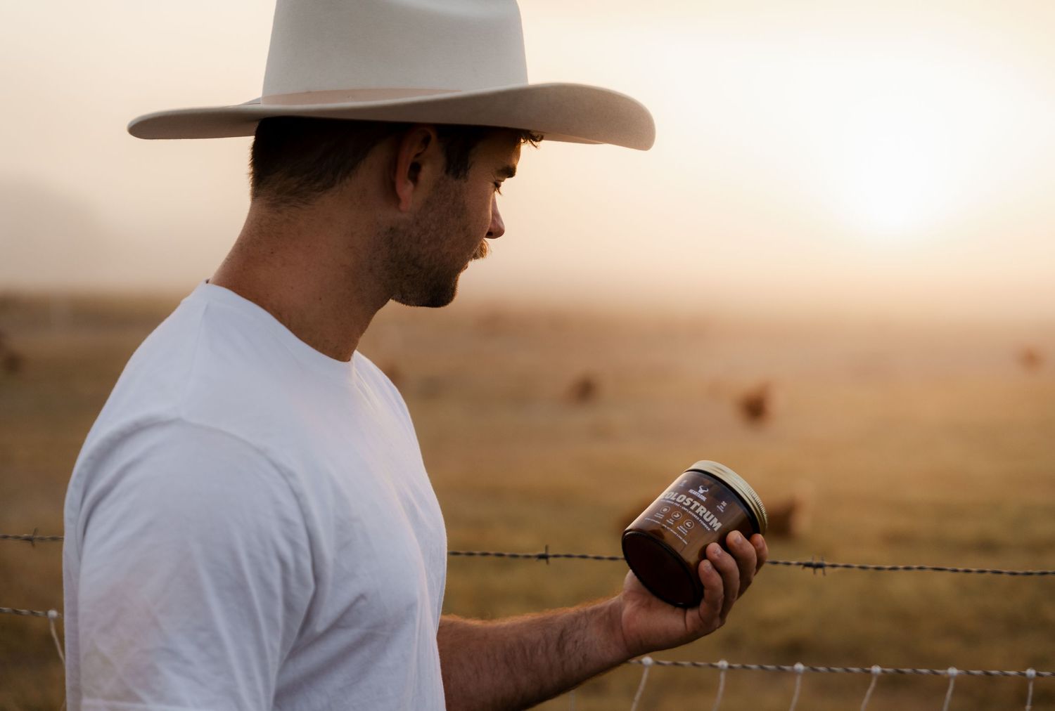 Man in white t-shirt and cowboy hat holding a jar of jam near a barbed wire fence at golden sunset.