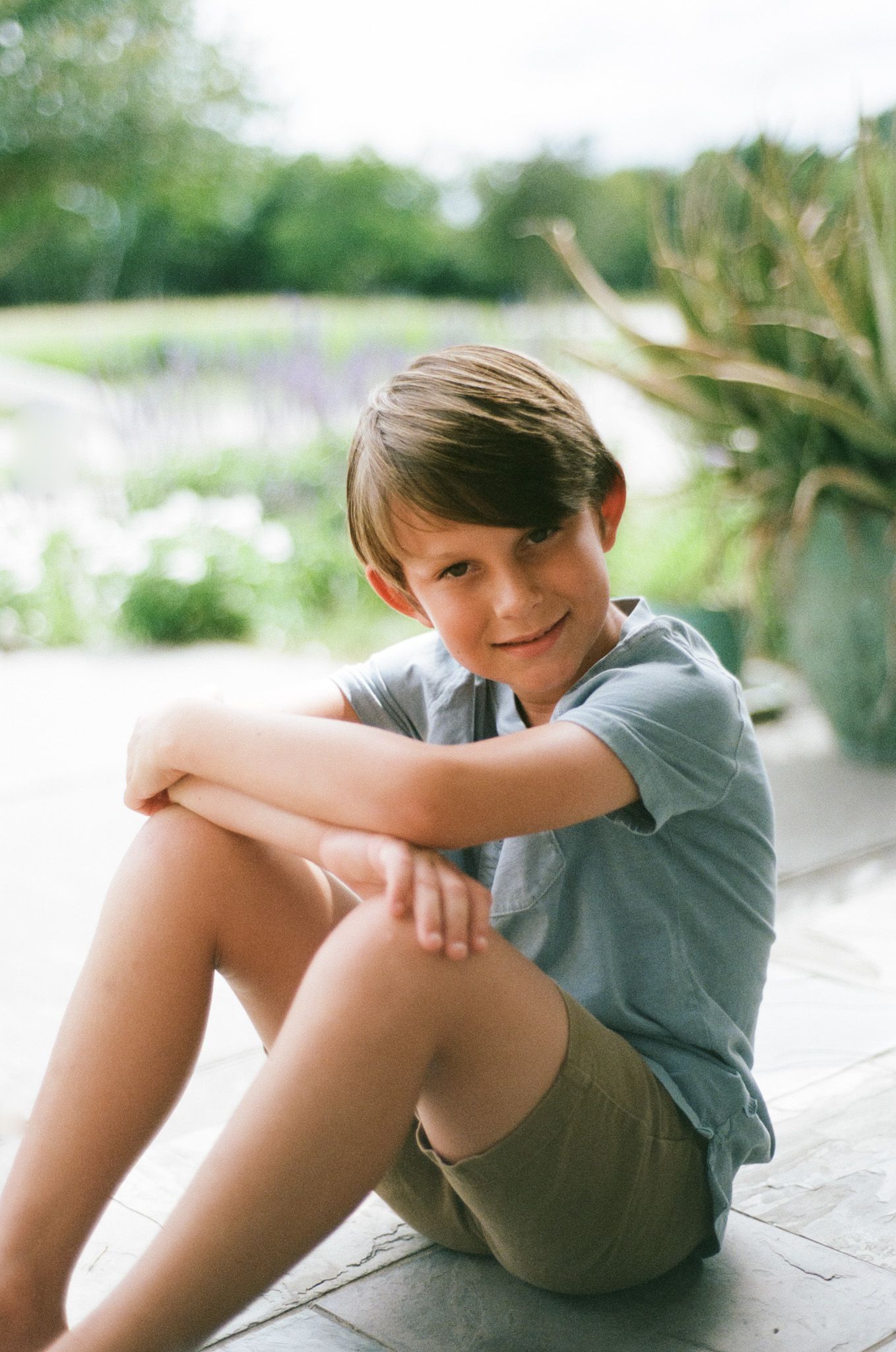 A smiling young boy in a blue shirt and khaki shorts sits relaxed on a patio surrounded by lush greenery.