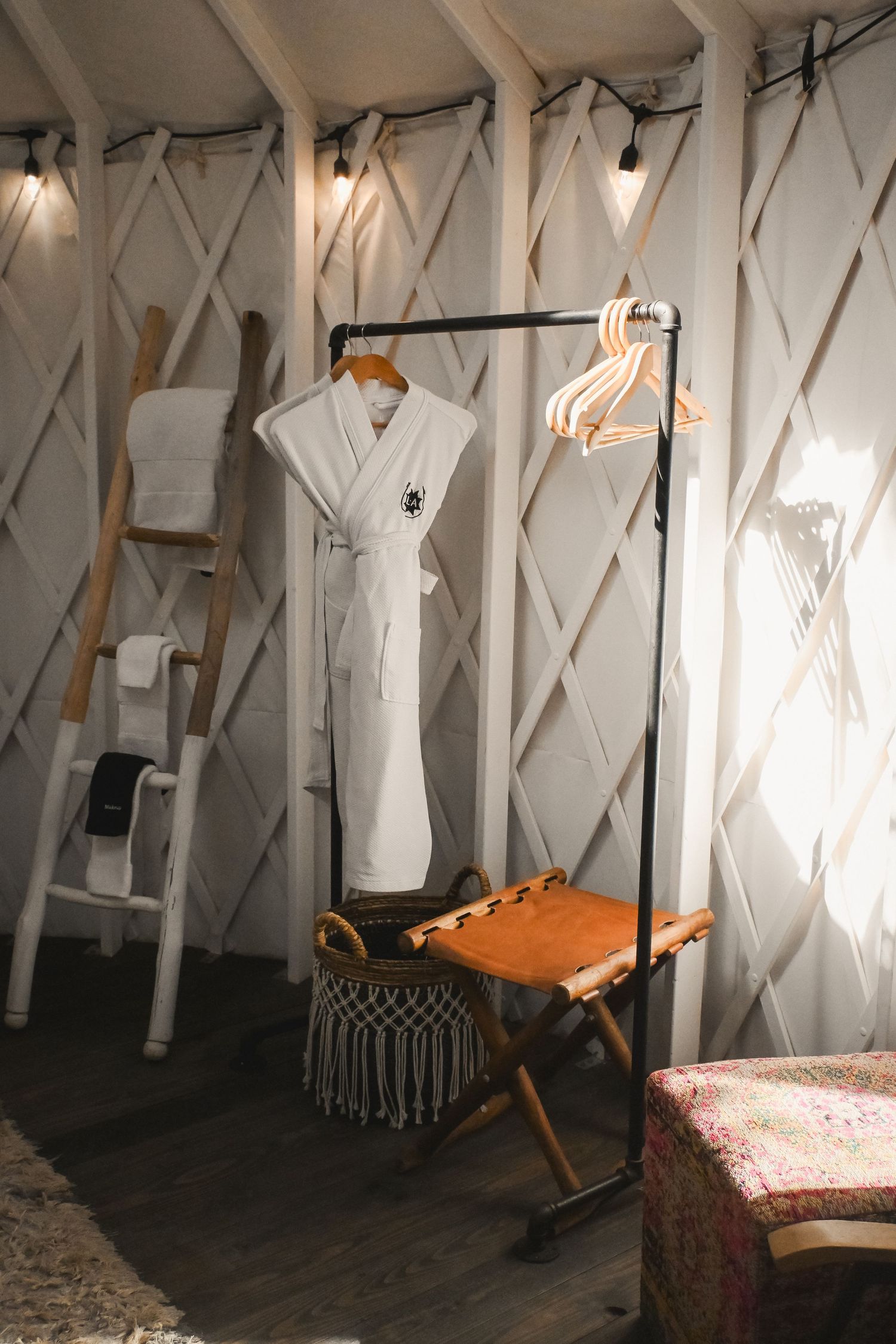 Yurt interior with a white bathrobe on a clothing rack, ladder shelf, and warm string lights.