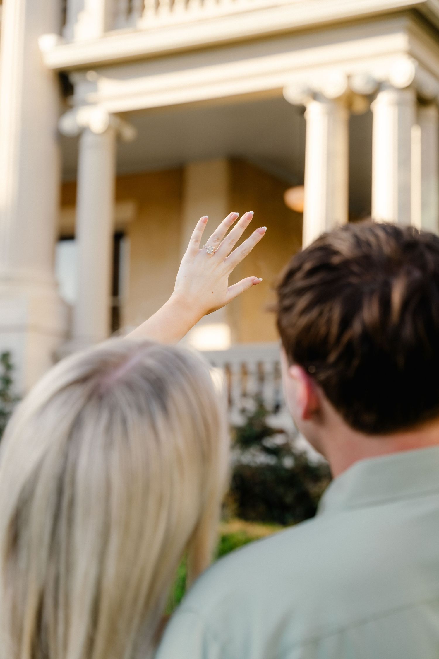 Engaged couple admiring a historic columned mansion, woman showing off her engagement ring.