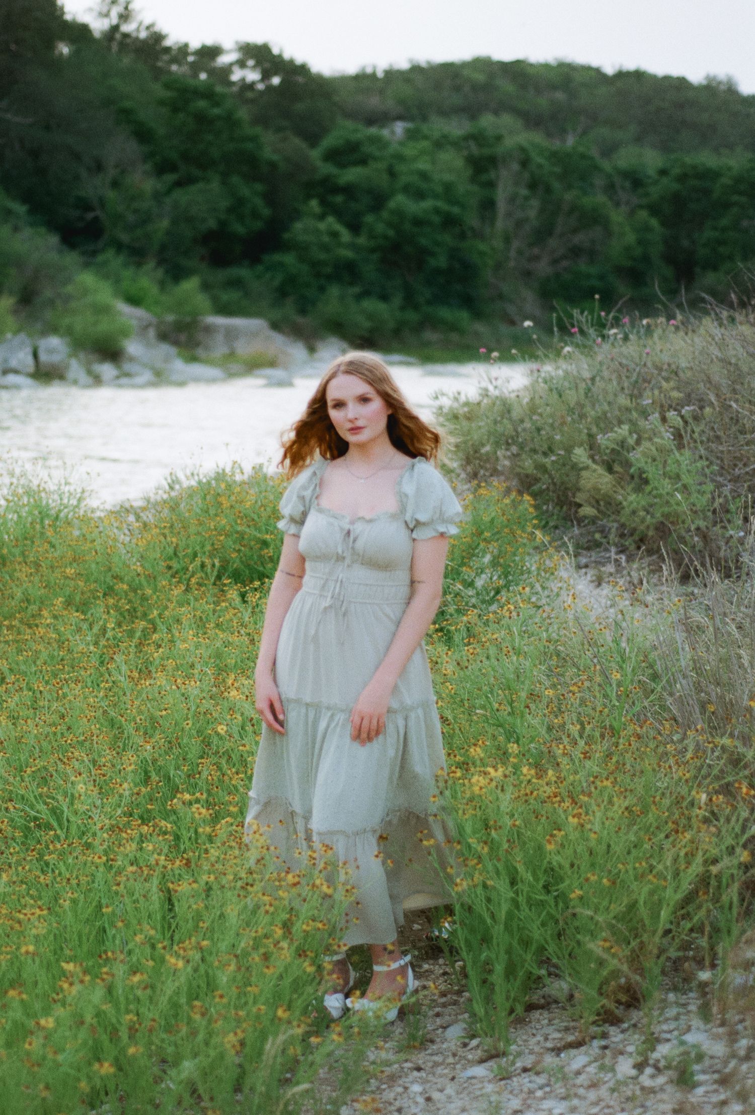 Young woman in sage green cottagecore midi dress standing among wildflowers by a river with lush green trees.