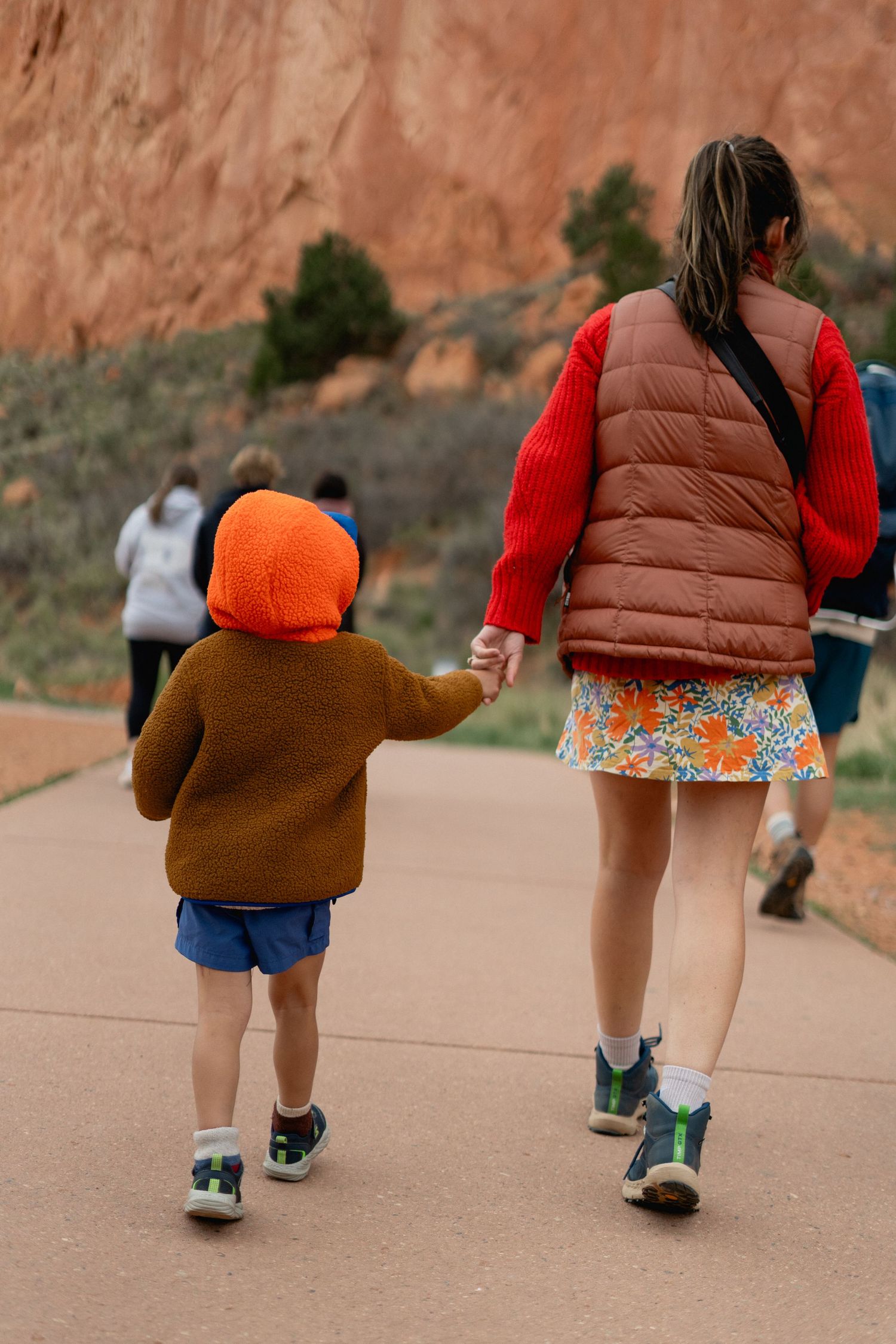 A woman and young child in an orange hat walk hand-in-hand on a paved path near red rock formations.