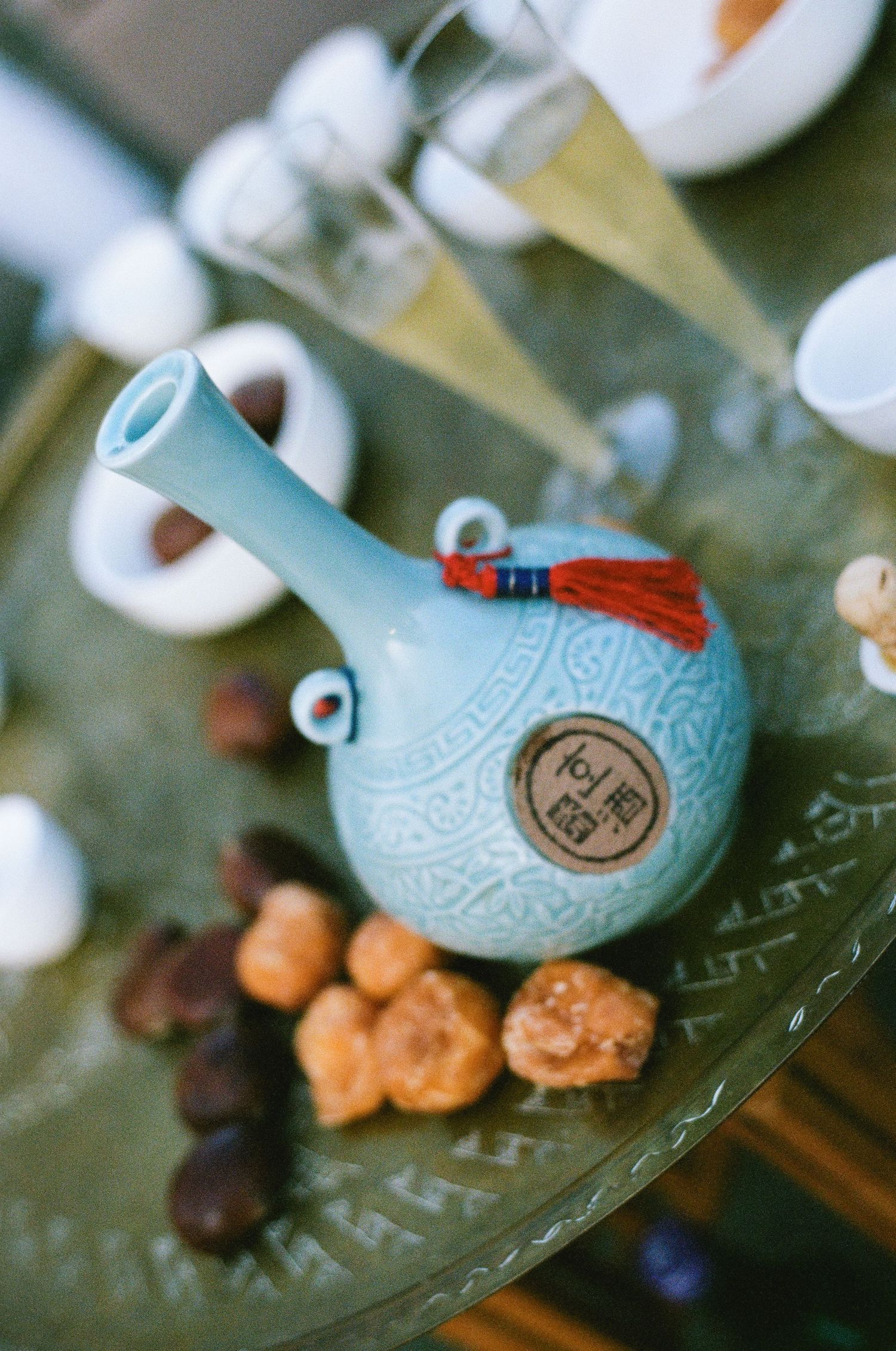 A light blue ceramic Korean tea vessel with red tassel sits on a decorative plate with dried fruits.