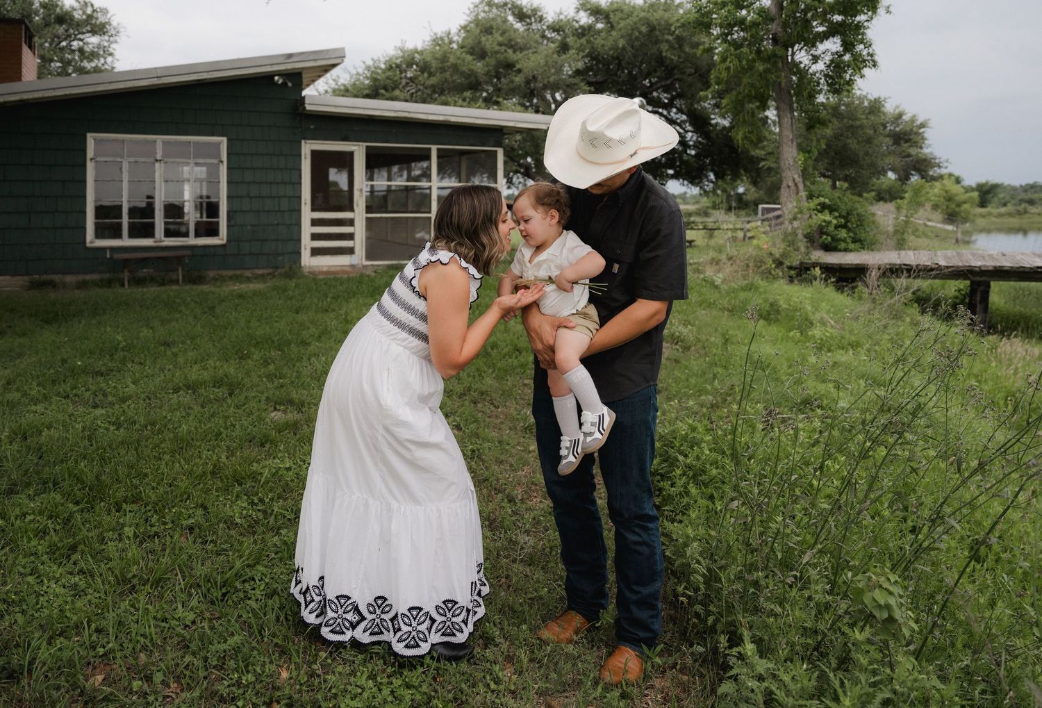 Young family with baby sharing a tender moment in a green yard outside a farmhouse.