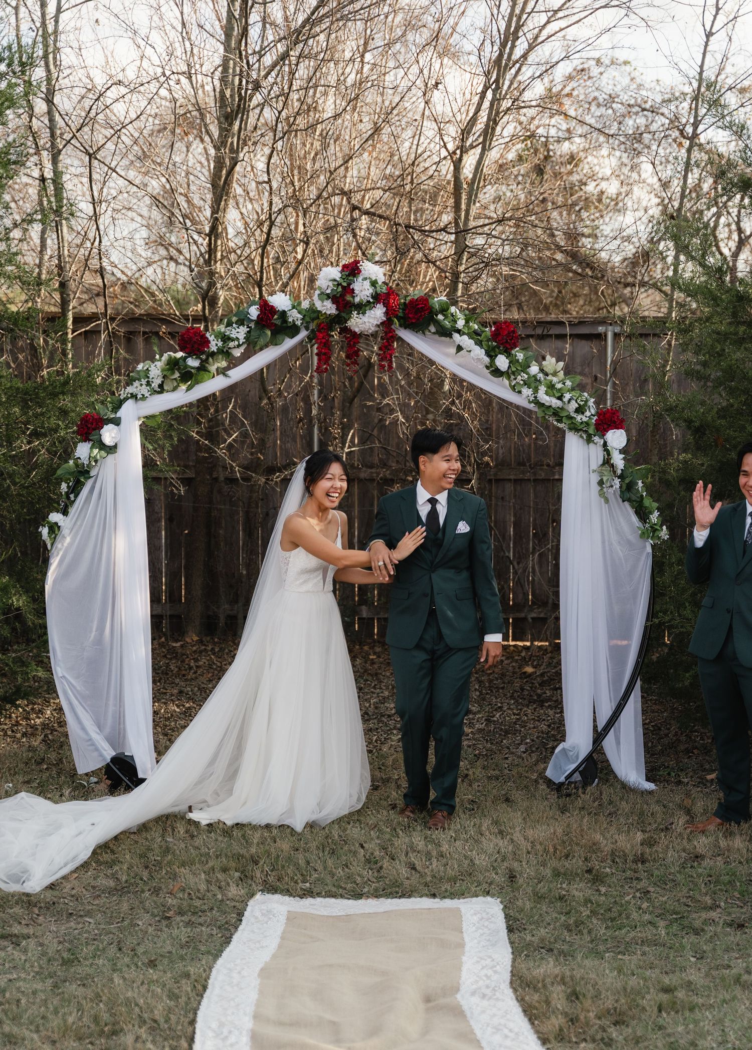 Smiling bride and groom stand under white draped arch adorned with red and white roses.