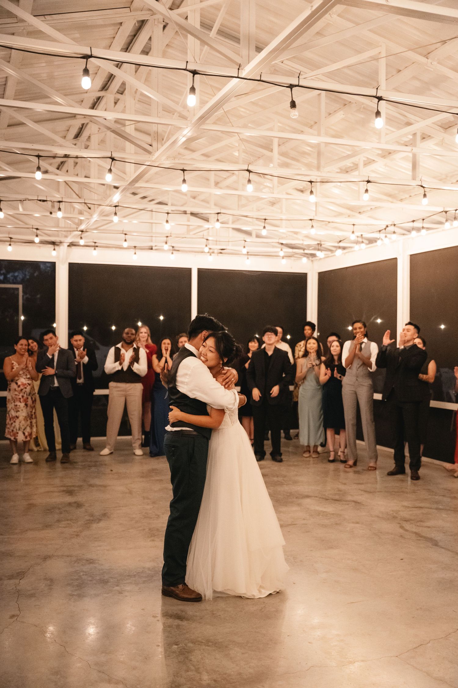 Bride and groom share first dance under string lights while wedding guests applaud in elegant venue.