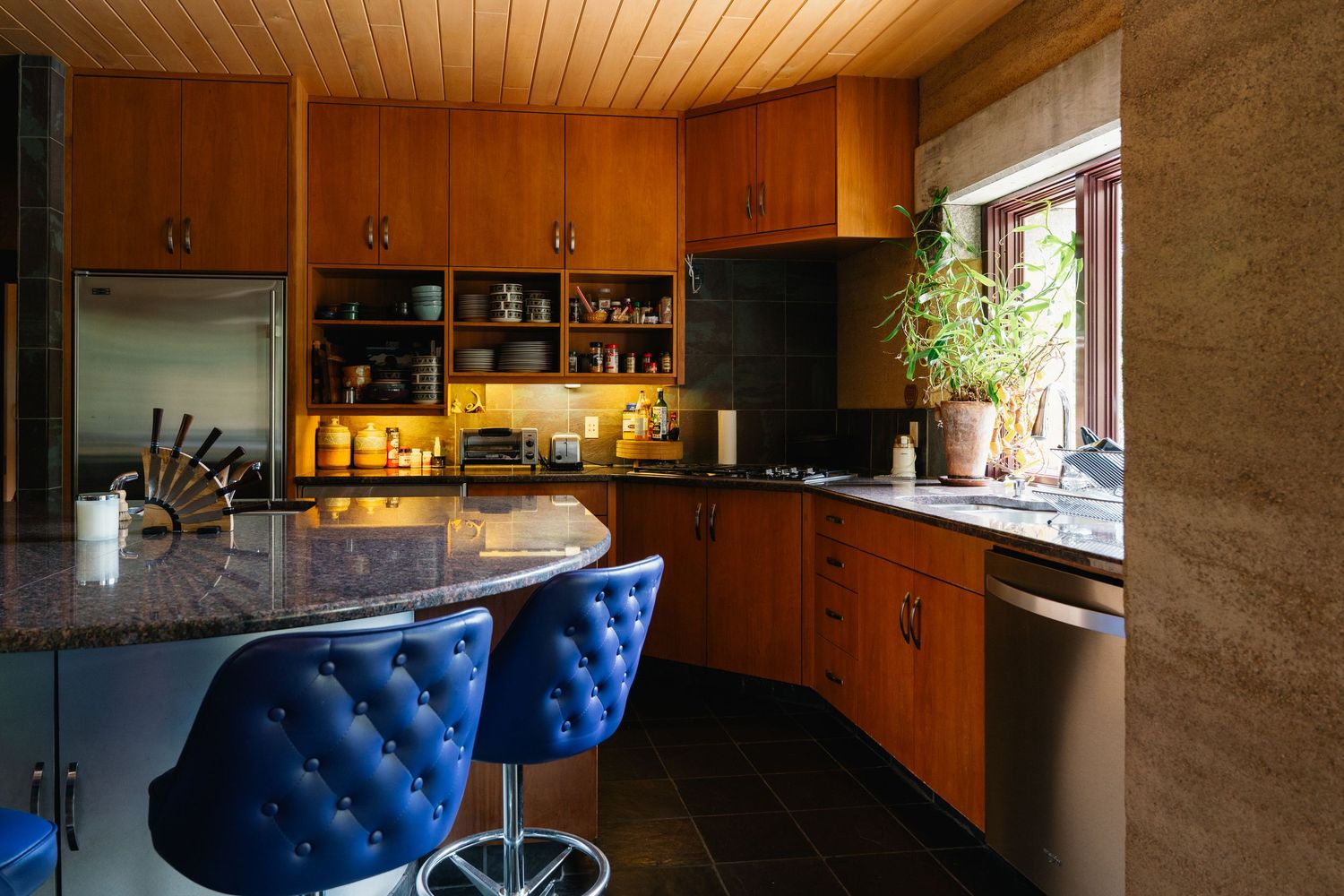 Mid-century modern kitchen with warm wood cabinets, granite island, blue bar stools, and wood plank ceiling.