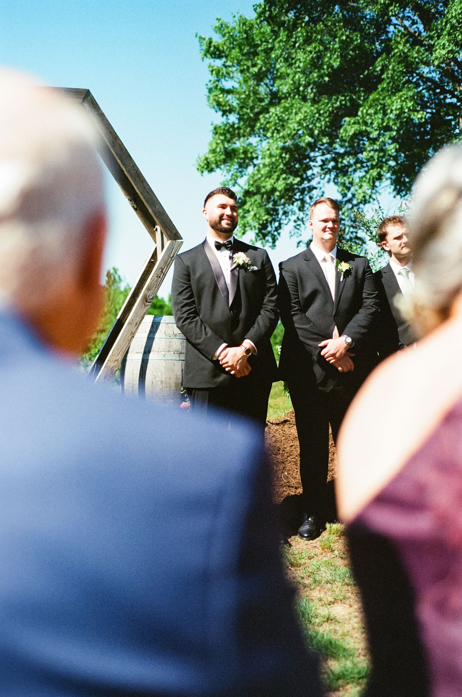 Smiling groom in black suit with boutonniere standing at outdoor wedding ceremony with guests.
