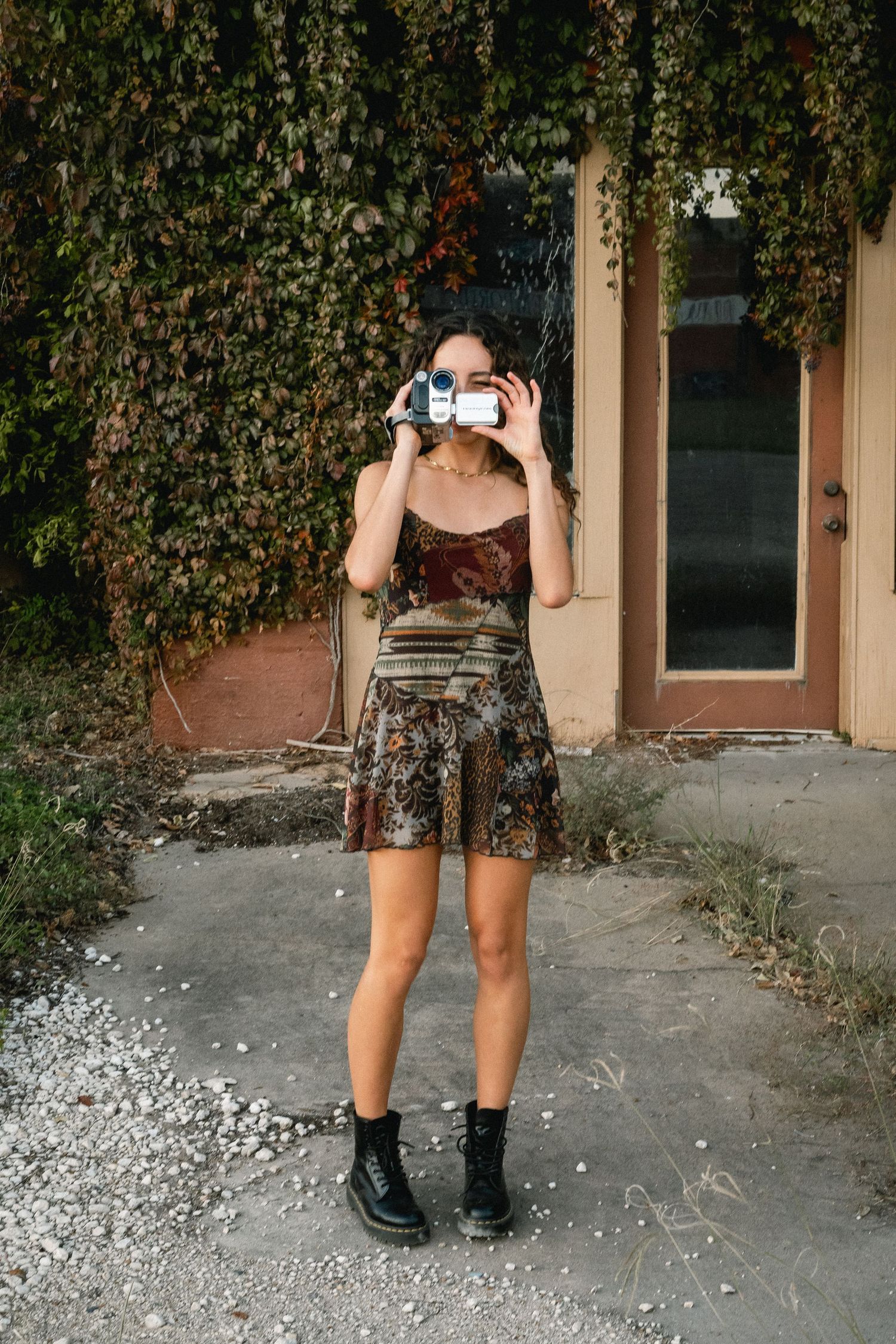 Young woman in brown velvet dress and black boots photographing with a camcorder outdoors near ivy-covered wall.