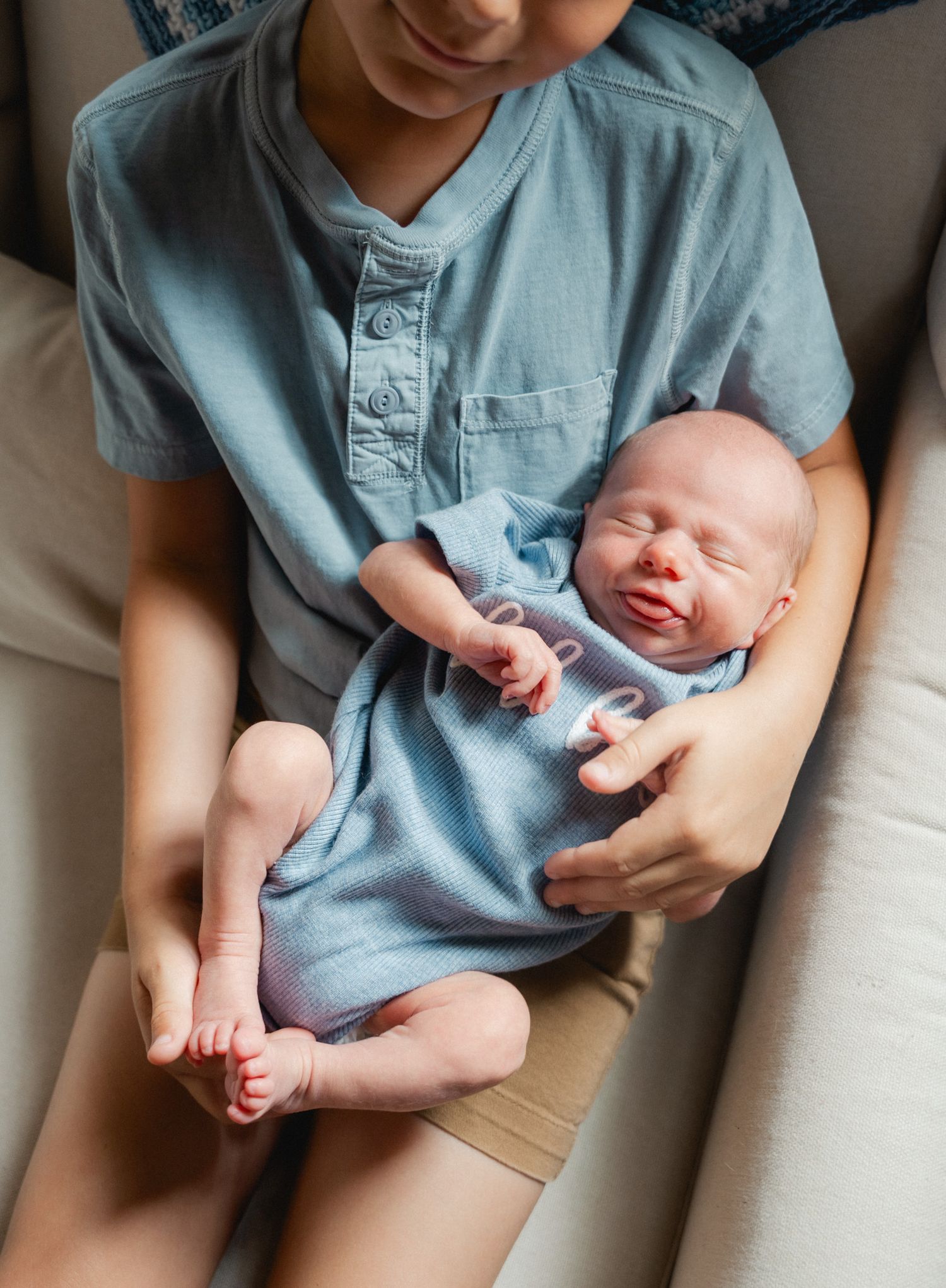 Older boy in blue shirt gently cradles sleeping newborn baby in his arms.