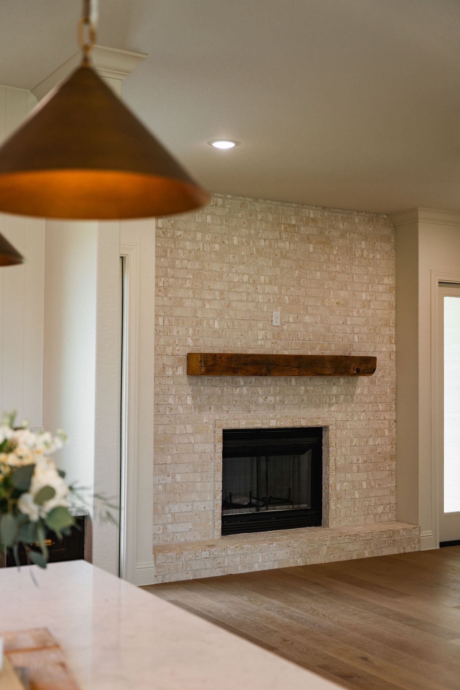 Neutral brick fireplace with rustic wood mantel in a modern home with hardwood floors and brass pendant light.
