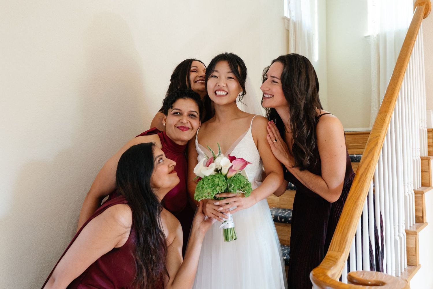 Bride holding bouquet laughs with bridesmaids in burgundy dresses on staircase before wedding ceremony.