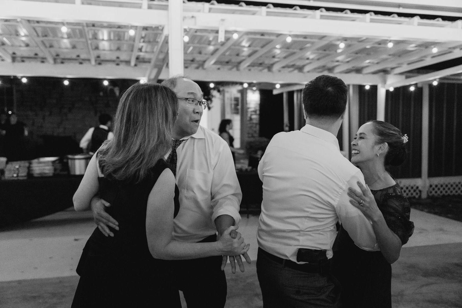 Black and white photo of couples slow dancing together at a wedding reception under string lights.