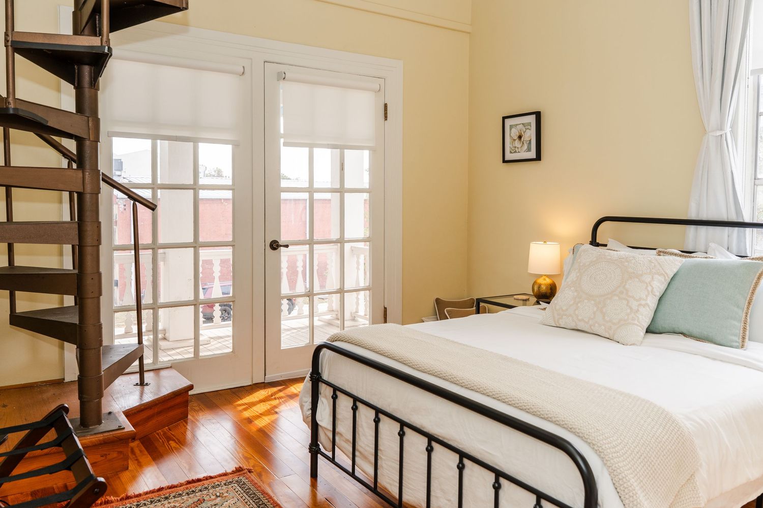 Charming bedroom with black iron bed frame, French doors leading to a balcony, spiral staircase, and hardwood floors.