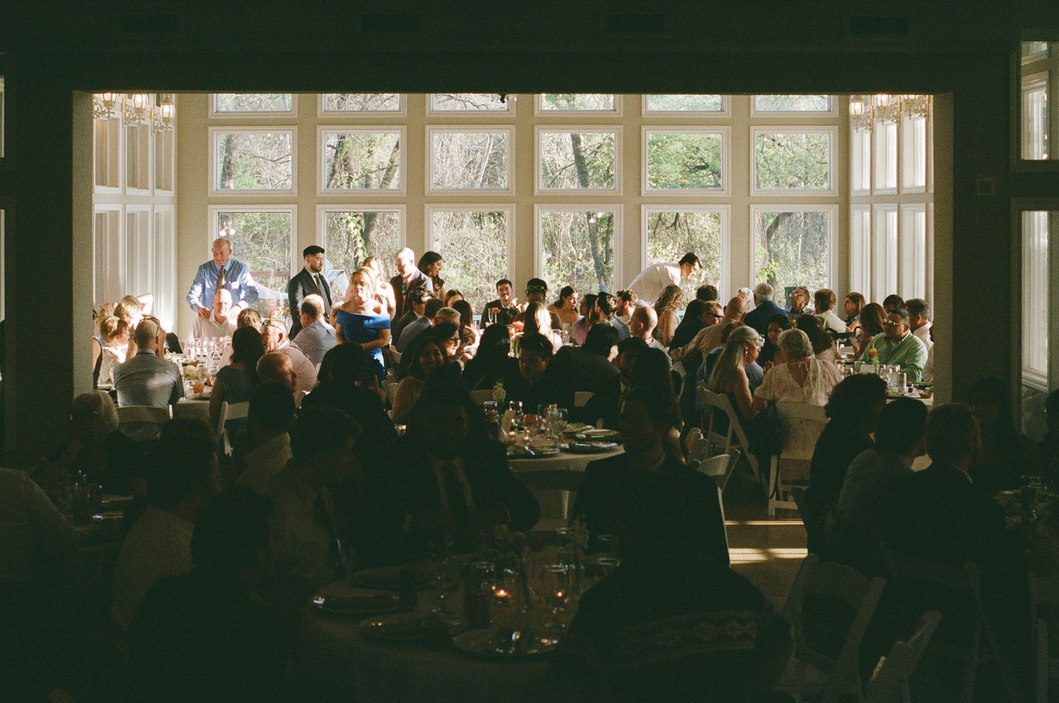 Crowded wedding reception in a sunlit venue with large windows, guests gathered around tables in elegant formal attire.