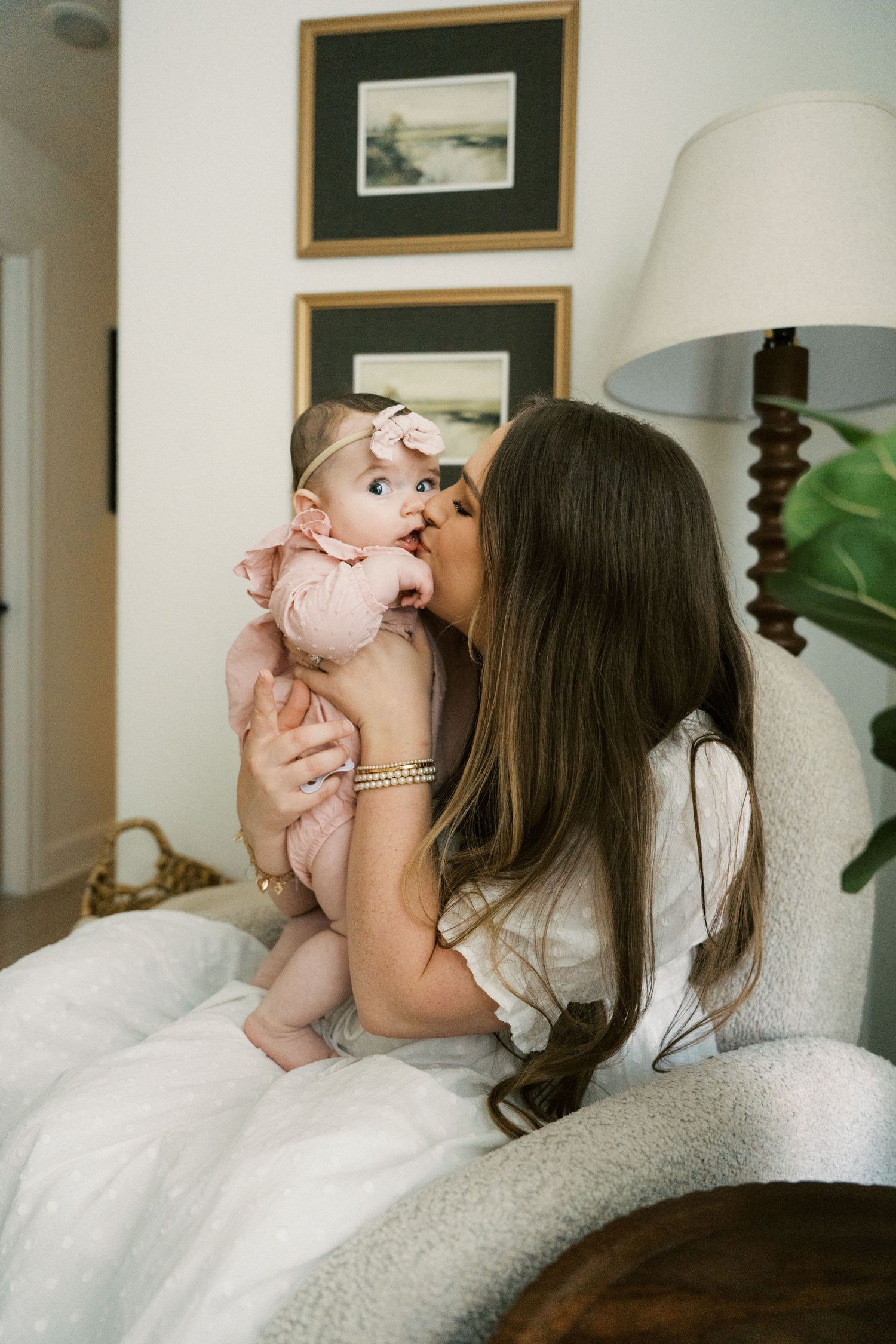 A mother with long brown hair kissing her baby girl dressed in pink on a white chair.