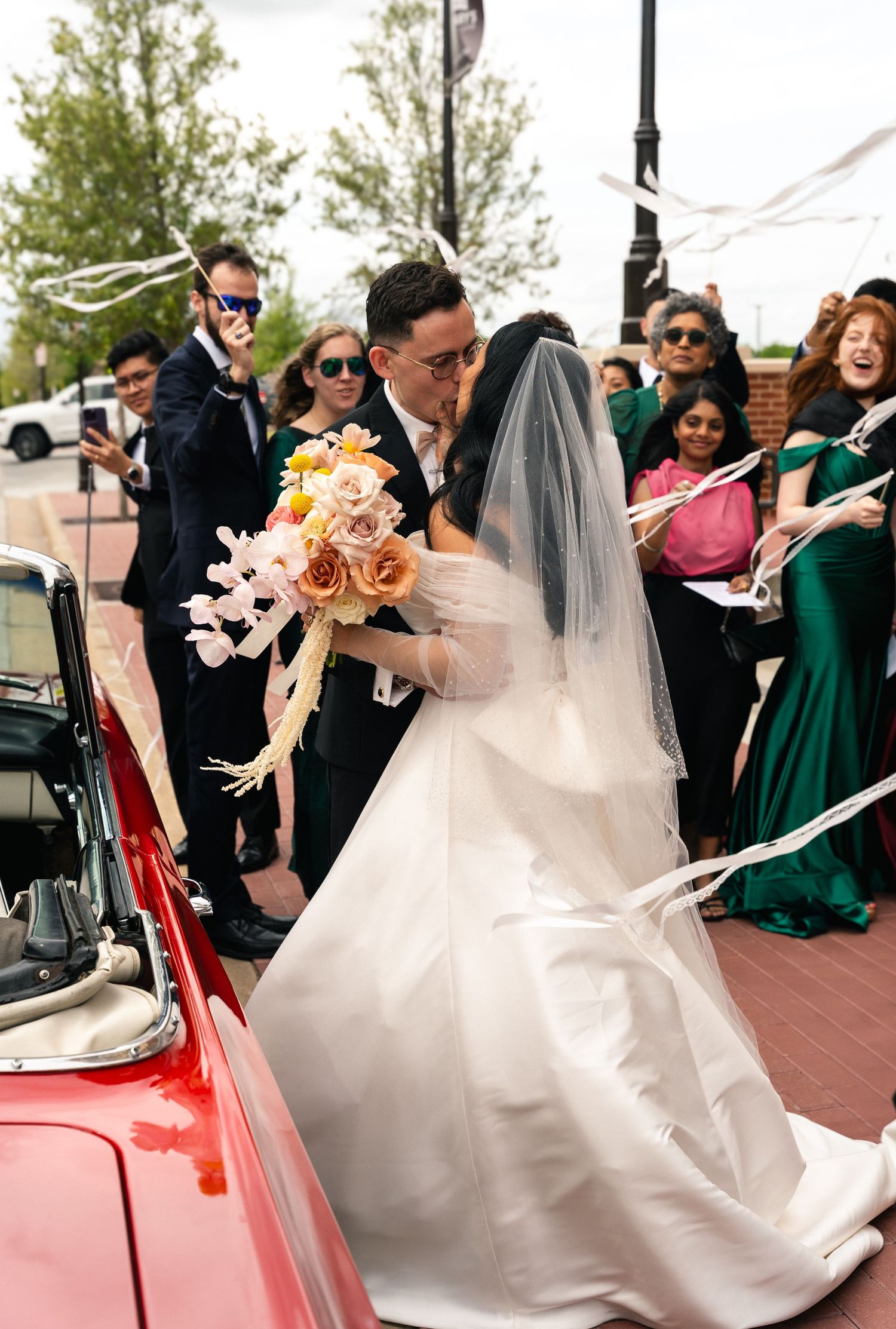 Newlyweds sharing a kiss beside a vintage red convertible surrounded by cheering wedding guests.